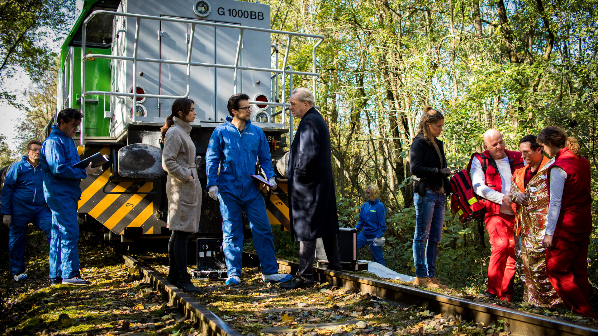 "SOKO München - Kain und Abel": Am Tatort steht ein Schienenfahrzeug auf Schienen im Wald Katharina Hahn (Bianca Hein), Dr. Weissenböck (Florian Odendahl) und Arthur Bauer (Gerd Silberbauer) stehen davor auf den Schienen und besprechen sich. Auf der einen Seite stehen Kollegen der Spusi (Komparsen), auf der anderen Seite steht Toni Bischoff (Amanda da Gloria) mit Sanitätern, die ein Opfer gerade in eine Überlebensfolie wickeln.