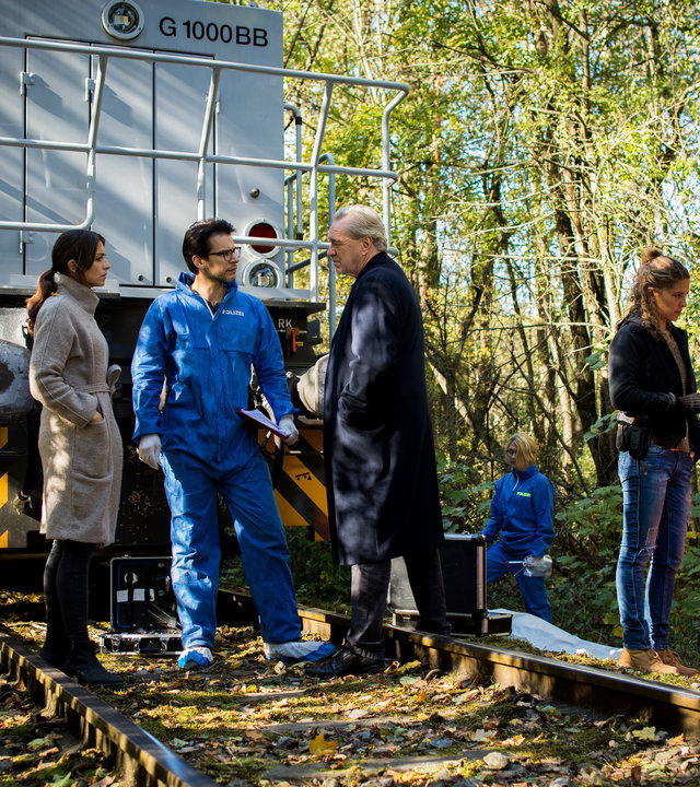 "SOKO München - Kain und Abel": Am Tatort steht ein Schienenfahrzeug auf Schienen im Wald Katharina Hahn (Bianca Hein), Dr. Weissenböck (Florian Odendahl) und Arthur Bauer (Gerd Silberbauer) stehen davor auf den Schienen und besprechen sich. Auf der einen Seite stehen Kollegen der Spusi (Komparsen), auf der anderen Seite steht Toni Bischoff (Amanda da Gloria) mit Sanitätern, die ein Opfer gerade in eine Überlebensfolie wickeln.