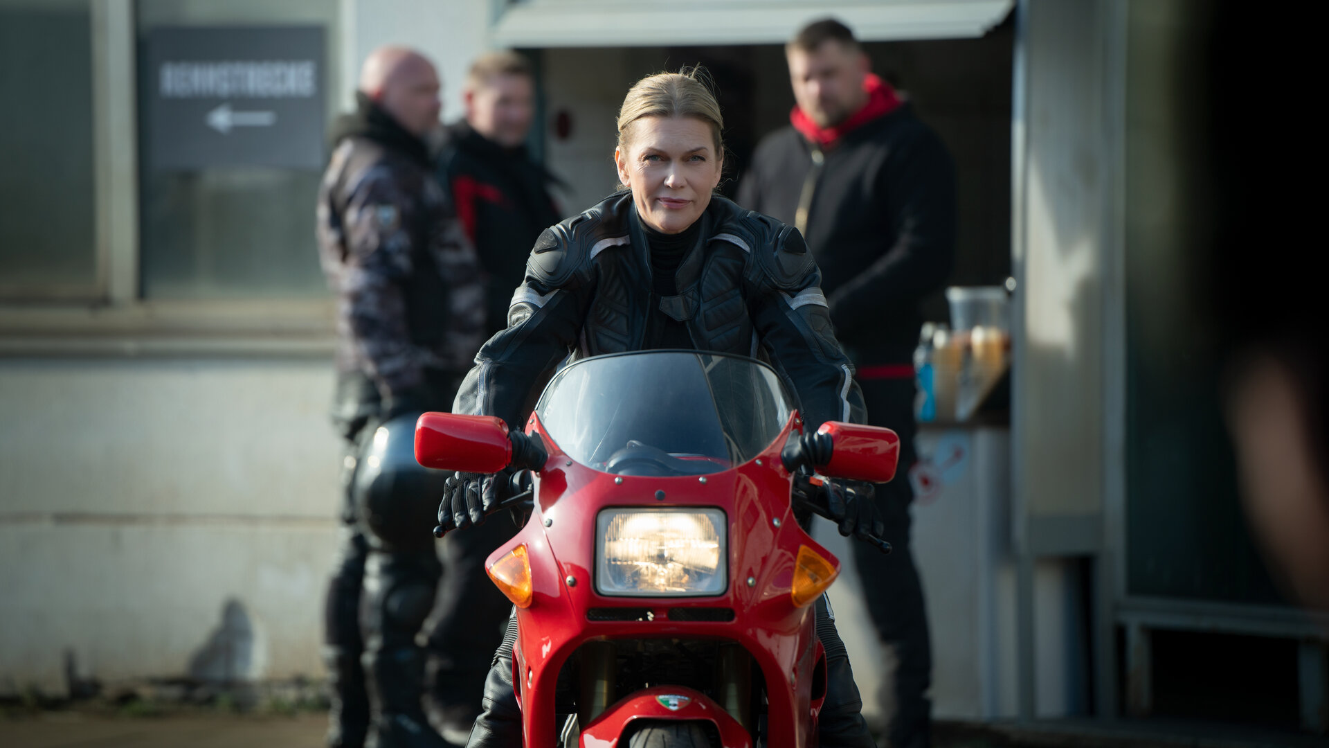 "Helen Dorn - Adrenalin": Helen Dorn sitzt auf einem Motorrad und fährt Richtung Kamera. Im Hintergrund, unscharf zu erkennen, stehen drei Männer vor einem Gebäude.
