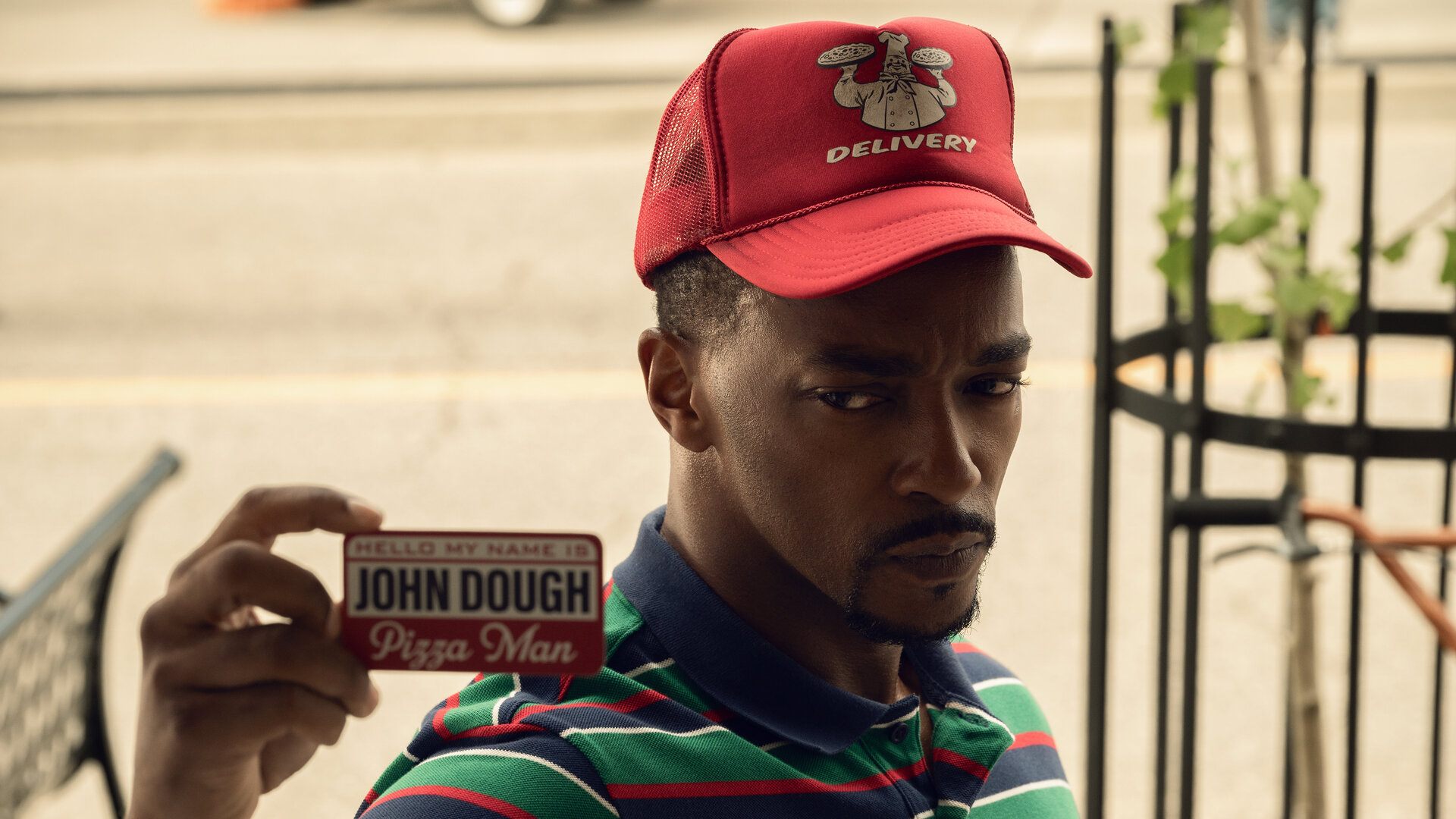 "Twisted Metal - PRSPRNT": John Doe (Anthony Mackie) steht bei Tag auf einer Straße. Er trägt ein bunt gestreiftes Shirt mit Kragen sowie eine rote Mütze mit weißem Pizzalieferdienst-Aufdruck. In der Hand hält er einen Dienstausweis mit dem Namen "John Dough", den er vor sich hält. Er blickt ernst nach vorn. Im Hintergrund befinden sich eine dunkelgraue Sitzbank und ein Baum.