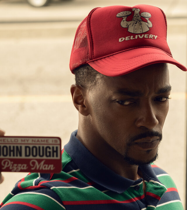 "Twisted Metal - PRSPRNT": John Doe (Anthony Mackie) steht bei Tag auf einer Straße. Er trägt ein bunt gestreiftes Shirt mit Kragen sowie eine rote Mütze mit weißem Pizzalieferdienst-Aufdruck. In der Hand hält er einen Dienstausweis mit dem Namen "John Dough", den er vor sich hält. Er blickt ernst nach vorn. Im Hintergrund befinden sich eine dunkelgraue Sitzbank und ein Baum.
