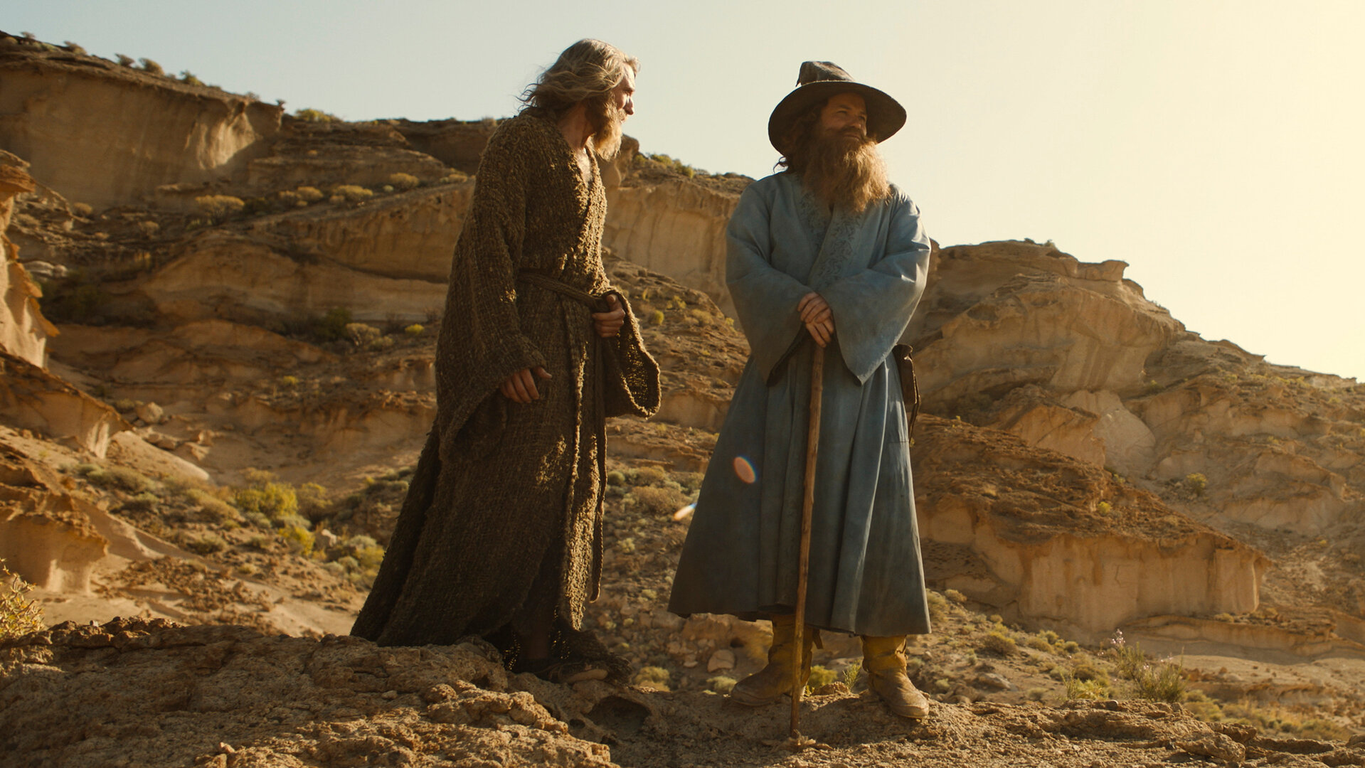 "Der Herr der Ringe: Die Ringe der Macht - Wo ist er?": Der Fremde (Daniel Weyman) und Tom Bombadil (Rory Kinnear) stehen nebeneinander in einer Wüstenlandschaft. Beide haben lange Bärte. Bombadil trägt einen Hut und stützt sich auf einen Stock. Der Fremde schaut in seine Richtung.