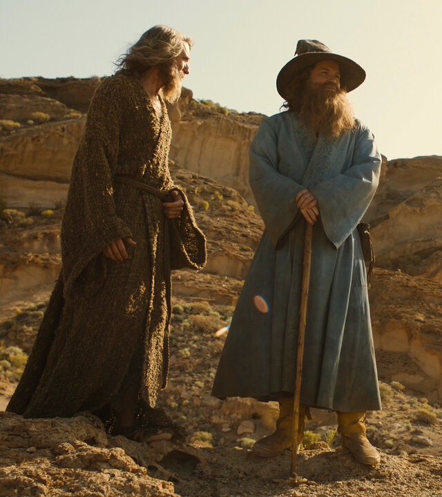 "Der Herr der Ringe: Die Ringe der Macht - Wo ist er?": Der Fremde (Daniel Weyman) und Tom Bombadil (Rory Kinnear) stehen nebeneinander in einer Wüstenlandschaft. Beide haben lange Bärte. Bombadil trägt einen Hut und stützt sich auf einen Stock. Der Fremde schaut in seine Richtung.