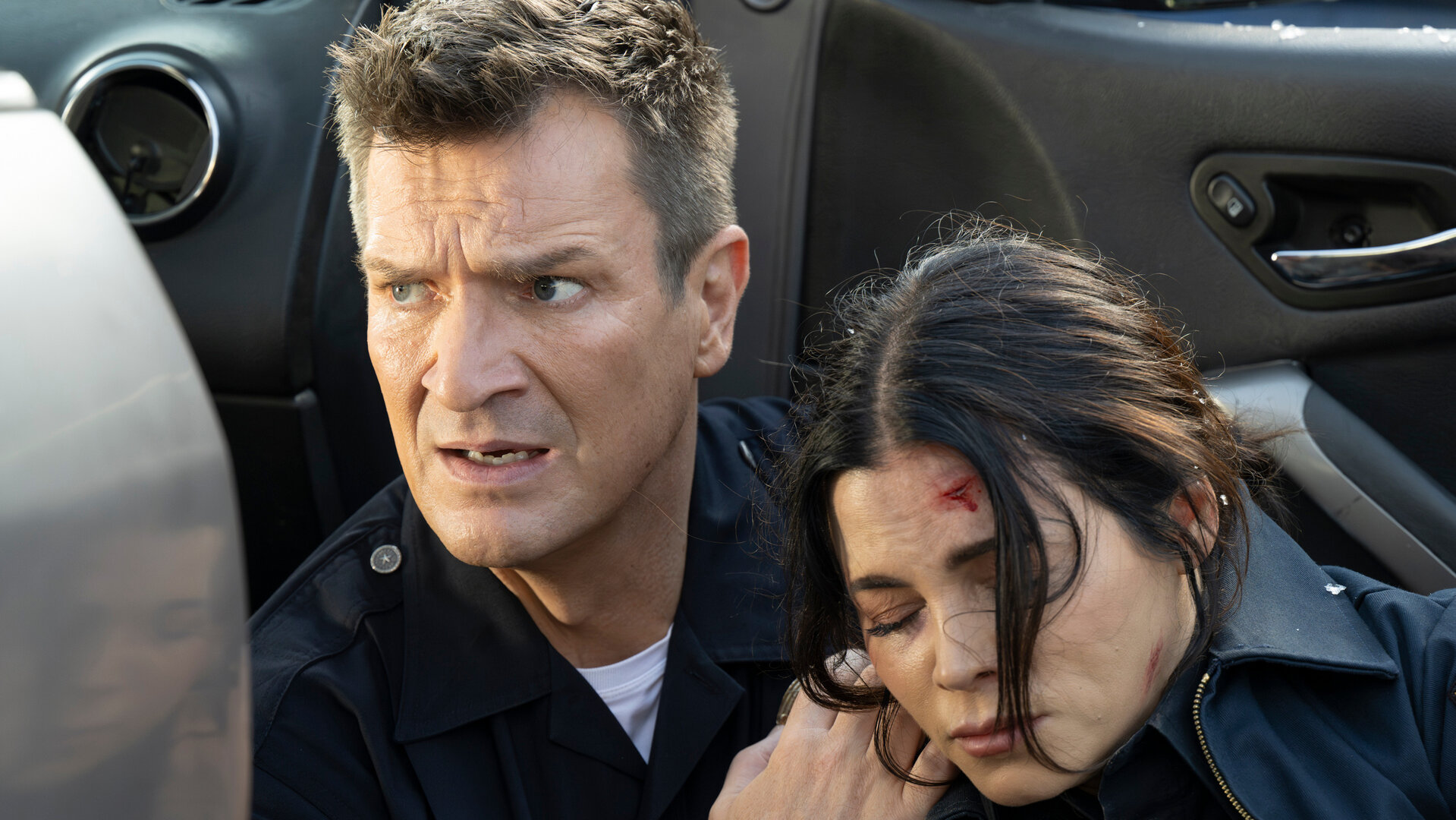 "The Rookie - Bis dass der Tod": Officer John Nolan (Nathan Fillion) und Bailey (Jenna Dewan) sitzen nebeneinander vor einer geöffneten Autotür. Nolan trägt eine dunkle Polizeiuniform, während Bailey eine dunkle Feuerwehruniform trägt. Er blickt mit nervösem Ausdruck zur Seite und hält Bailey an den Schultern fest. Bailey hat die Augen geschlossen und hat eine Platzwunde an der Stirn.
