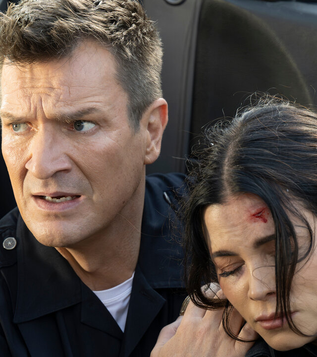 "The Rookie - Bis dass der Tod": Officer John Nolan (Nathan Fillion) und Bailey (Jenna Dewan) sitzen nebeneinander vor einer geöffneten Autotür. Nolan trägt eine dunkle Polizeiuniform, während Bailey eine dunkle Feuerwehruniform trägt. Er blickt mit nervösem Ausdruck zur Seite und hält Bailey an den Schultern fest. Bailey hat die Augen geschlossen und hat eine Platzwunde an der Stirn.