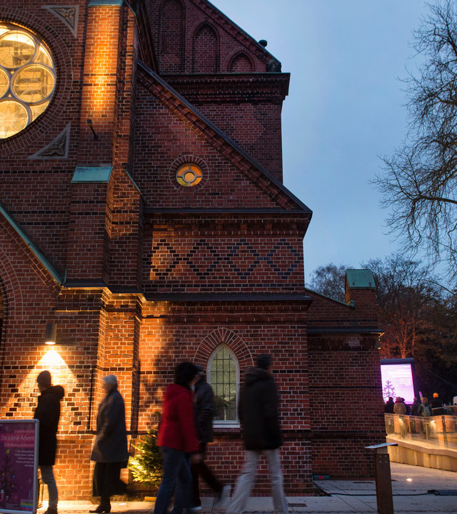 "Evangelischer Gottesdienst - Mehr als du erwartest": Außenansicht der St. Nicolaus Kirche im winterlich-abendlichen Hamburg-Alsterdorf: Sie ist aus rotem Backstein erbaut, vor ihrem Eingang laufen Passanten, neben der Kirche steht ein blätterloser Baum. Aus dem Innern der Kirche heraus scheint ein warmes Licht durch ein rundes Fenster oberhalb der geöffneten Eingangstür.