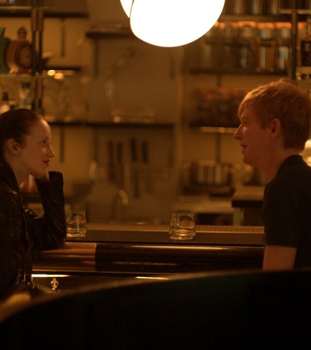 "Alice & Jack - Blind Date": In einer Kneipe sitzen Alice (Andrea Riseborough) und Jack (Domhnall Gleeson) nebeneinander an der Theke und schauen sich an. Sie reden miteinander. Hinter ihnen stehen viele, unterschiedliche Flaschen im beleuchteten Regal. Im Hintergrund, nur unscharf zu erkennen, sitzt eine weitere Person (Komparse).