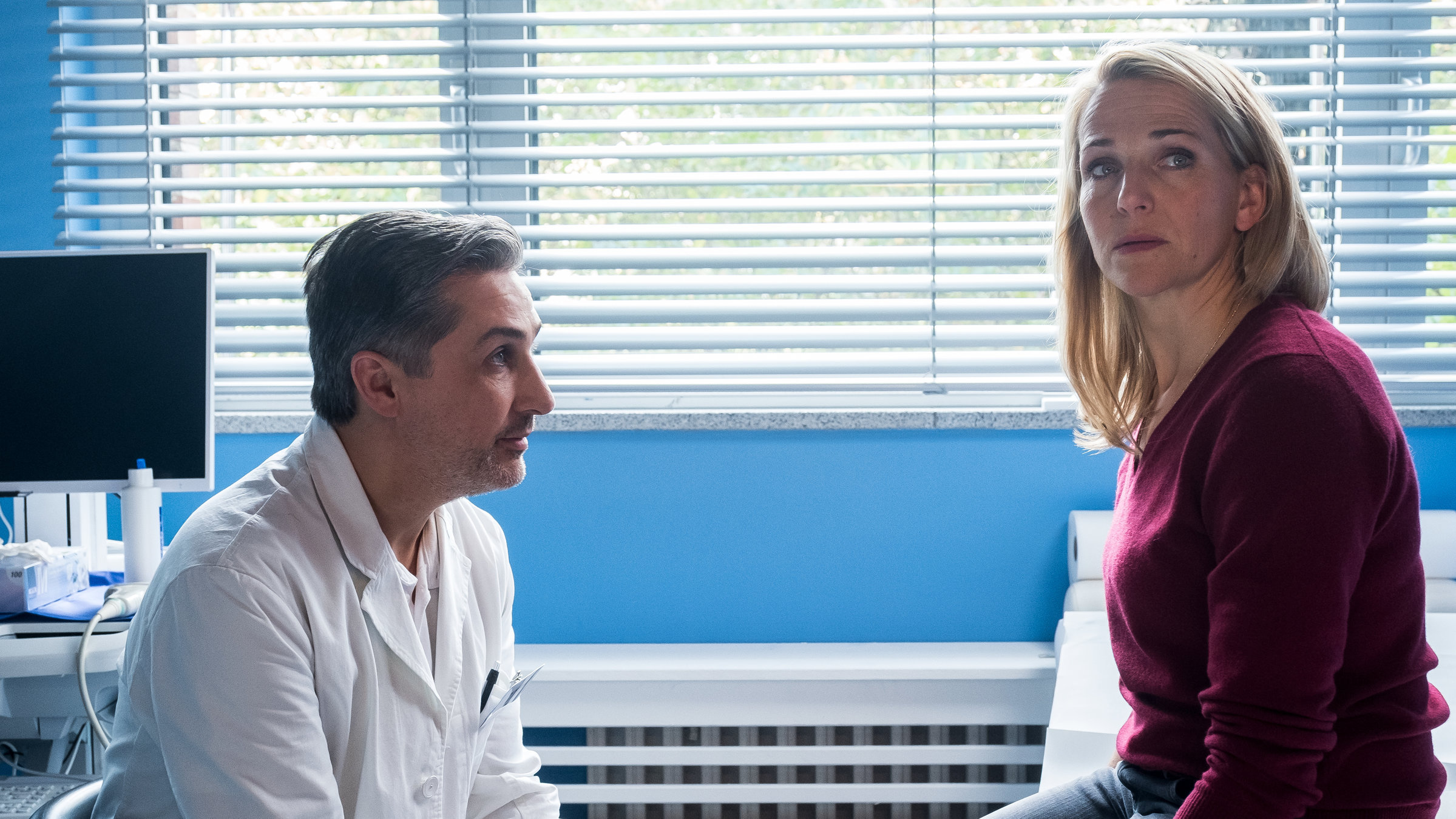 "Fritzie - Der Himmel muss warten - Was wirklich zählt": Anästhesist Anton Herzog (Rafi Guessous) sitzt in seinem Arztzimmer der besorgten Fritzie (Tanja Wedhorn) gegenüber. Sie sitzt auf einer Liege, er auf einem Stuhl.