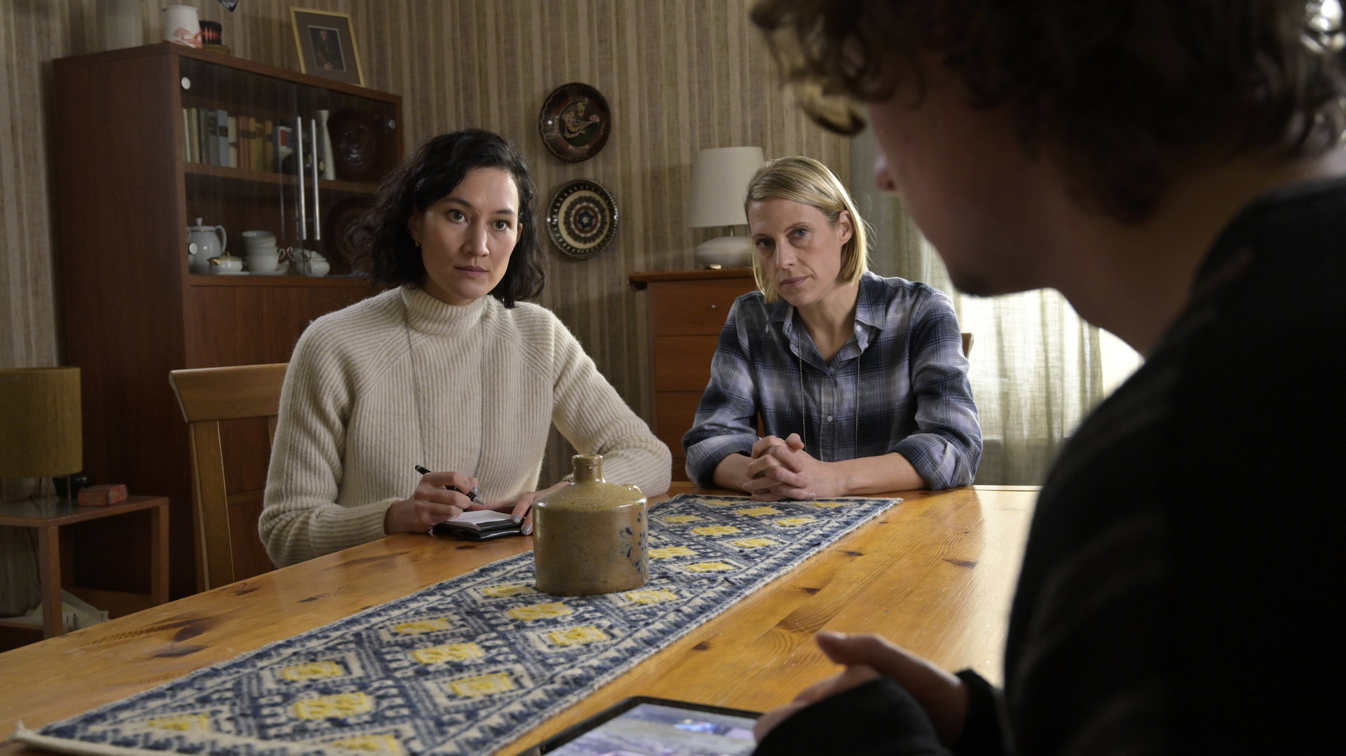"SOKO Potsdam - Fremder Vertrauter": An einem Esszimmertisch aus Holz sitzen die beiden  Kommissarinnen Pauline Hobrecht (Agnes Decker) und Tamara Meurer (Anja Pahl). Gegenüber von ihnen, mit dem Rücken zur Kamera, sitzt Tim Demandt (Jean Luc Caputo). Mit ernsten Blicken, sehen sie Tim Demandt an. In der rechten Hand hält Pauline Hobrecht einen Stift, vor ihr auf dem Tisch liegt ein Notizblock. Tim Demandt hat ein Tablet vor sich auf dem Tisch liegen.