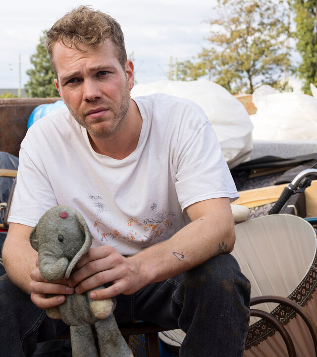 "Like a Loser: Ernste Sorge um Bobo": Julian (Ben Münchow) sitzt in einem Container voller Sperrmüll, umgeben von alten Möbeln, schwarzen Säcken und einem Fahrrad. Er hält den abgenutzten blauen Stoffelefanten Bobo fest in den Händen.
