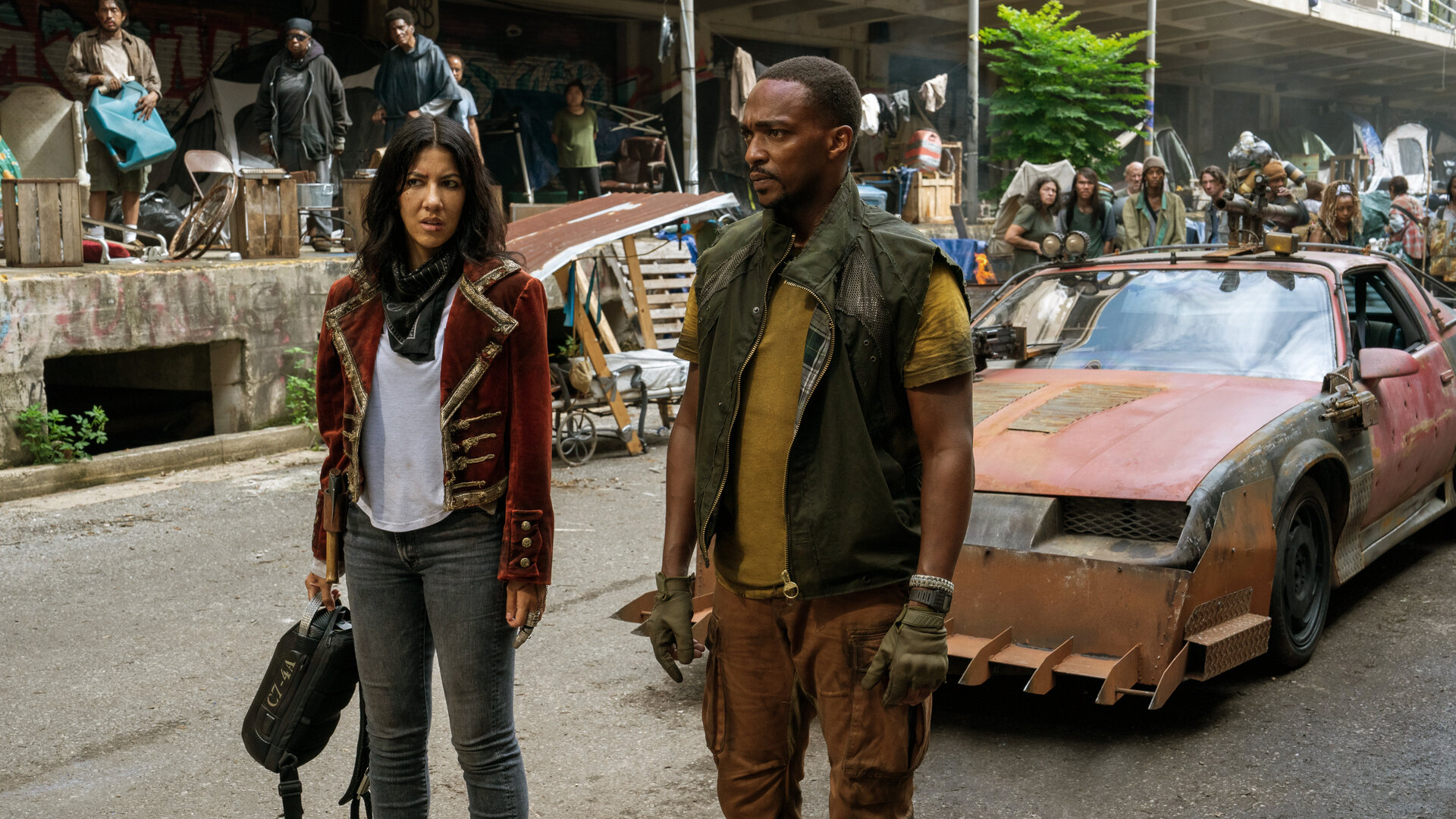 "Twisted Metal - SHNGRLA": John (Anthony Mackie) und Quiet (Stephanie Beatriz) stehen nebeneinander auf einer belebten Straße und blicken skeptisch in dieselbe Richtung. Quiet hält einen schwarzen Rucksack in der Hand. Hinter ihnen steht ein stark modifiziertes, orangefarbenes Auto, während im Hintergrund viele Personen (Komparsen) die beiden beobachten.