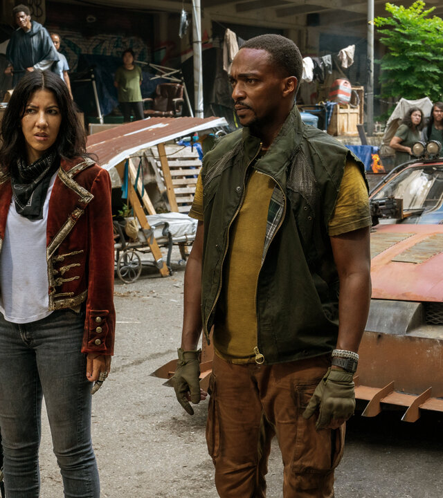 "Twisted Metal - SHNGRLA": John (Anthony Mackie) und Quiet (Stephanie Beatriz) stehen nebeneinander auf einer belebten Straße und blicken skeptisch in dieselbe Richtung. Quiet hält einen schwarzen Rucksack in der Hand. Hinter ihnen steht ein stark modifiziertes, orangefarbenes Auto, während im Hintergrund viele Personen (Komparsen) die beiden beobachten.
