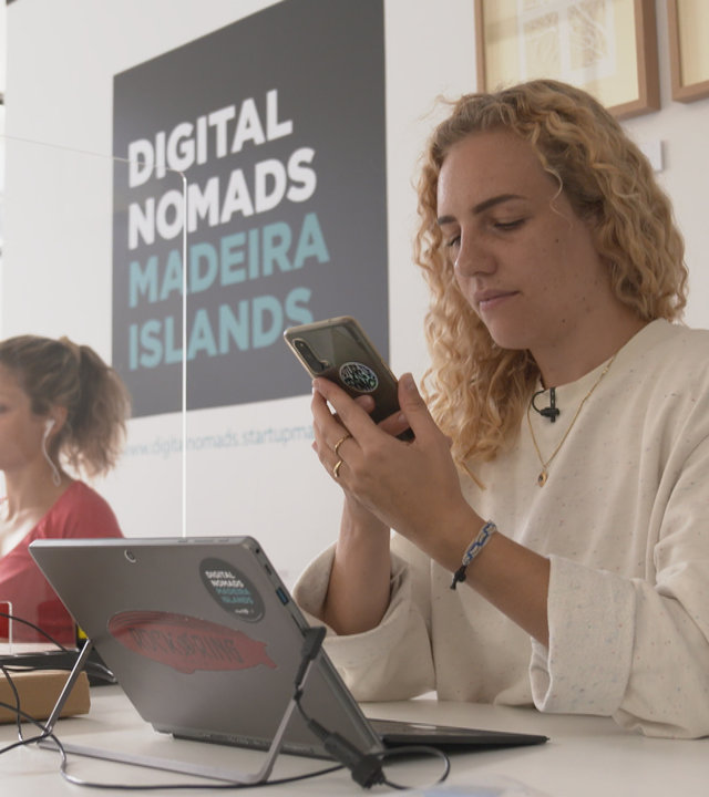 "Terra Xpress - Besser arbeiten - besser leben": Zwei Frauen sitzen an einem Schreibtisch und arbeiten am Laptop. Die blonde Frau im Vordergrund, Merle Makobe, um die es geht, hält ein Handy in der Hand. Im Hintergrund auf der Wand steht geschrieben: Digital Nomads Madeira Islands. Übersetzung: Digitale Nomaden der Insel Madeira.