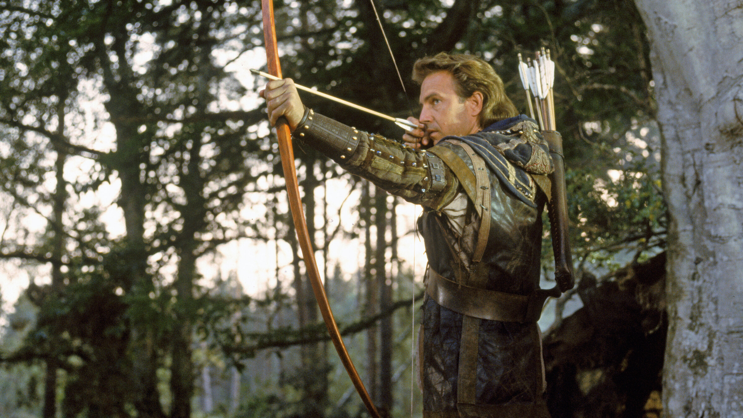 „Robin Hood – König der Diebe“: Robin Hood (Kevin Costner), in typischer, lederner Kluft und umgeben von Bäumen, zielt mit Pfeil und Bogen auf einen nicht sichtbaren Widersacher.