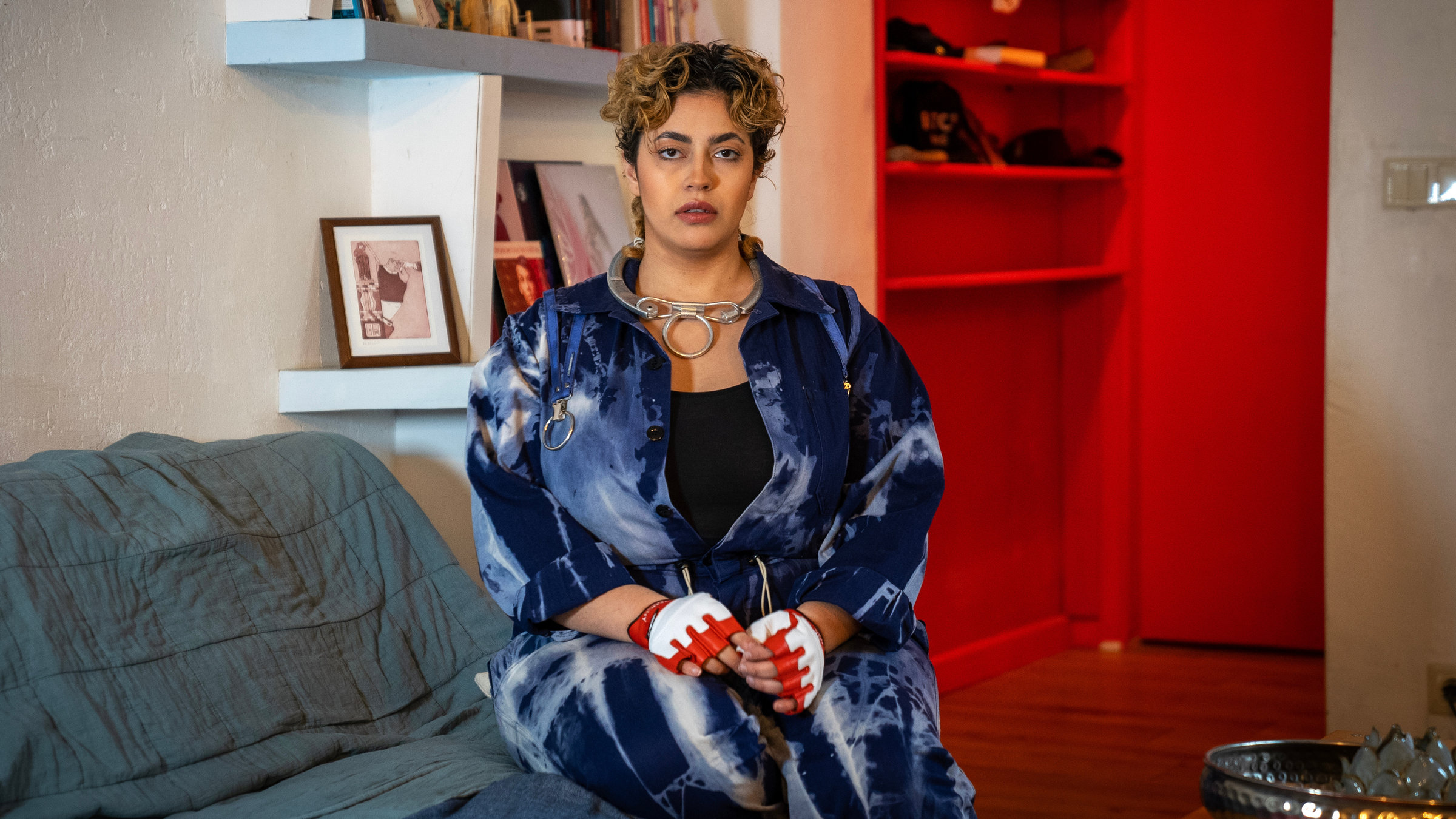 "Frau. Leben. Freiheit?": Frau mit einem Halsschmuck in Form einer Handschelle und rot-weißen Schutz-Handschuhe sitzt auf Sofakante vor Bücherregal und guckt frontal in die Kamera.