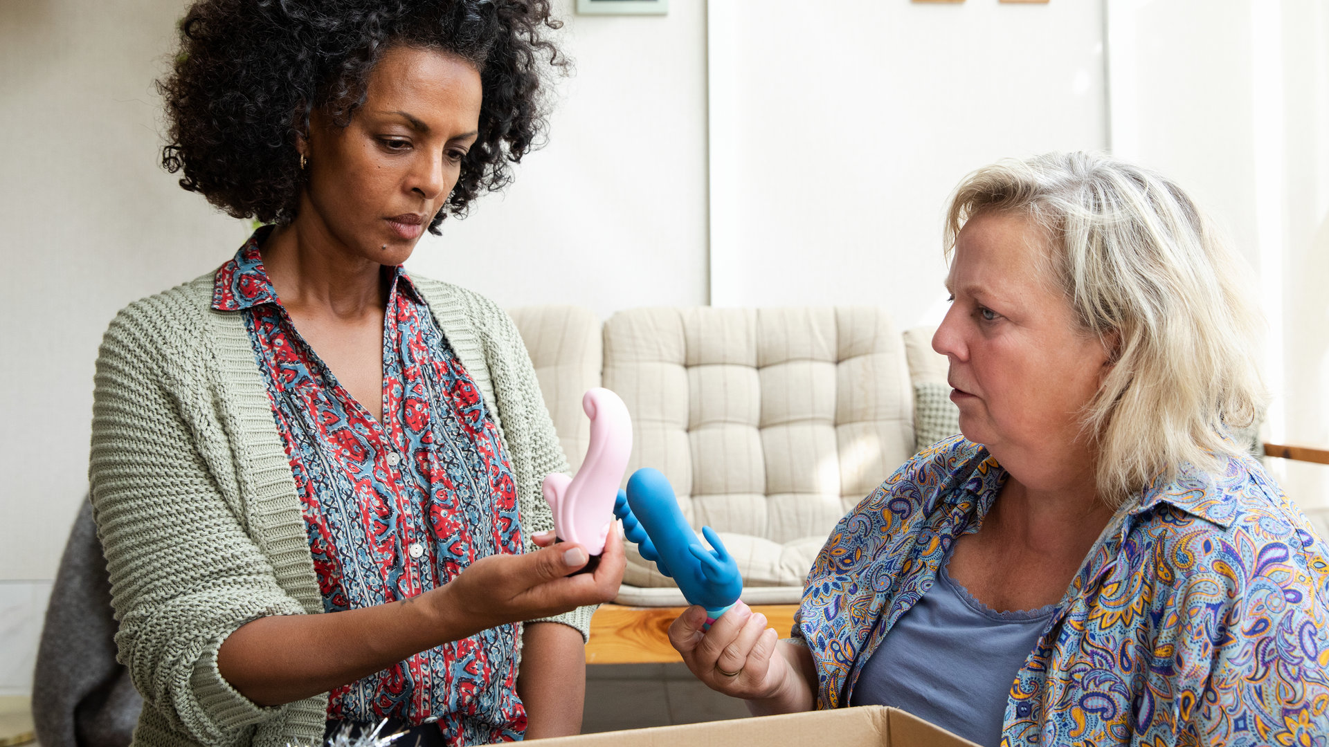 "I don't work here - Partykeller": Lemlem (Dennenesch Zoudé) und Heidi (Gabriela Maria Schmeide) sitzen im Wohnzimmer auf dem Boden vor einer geöffneten Kiste. Heidi hat einen blauen Vibrator in der Hand und zeigt ihn Lemlem, während diese einen rosa Vibrator in der Hand hält und ihn betrachtet.