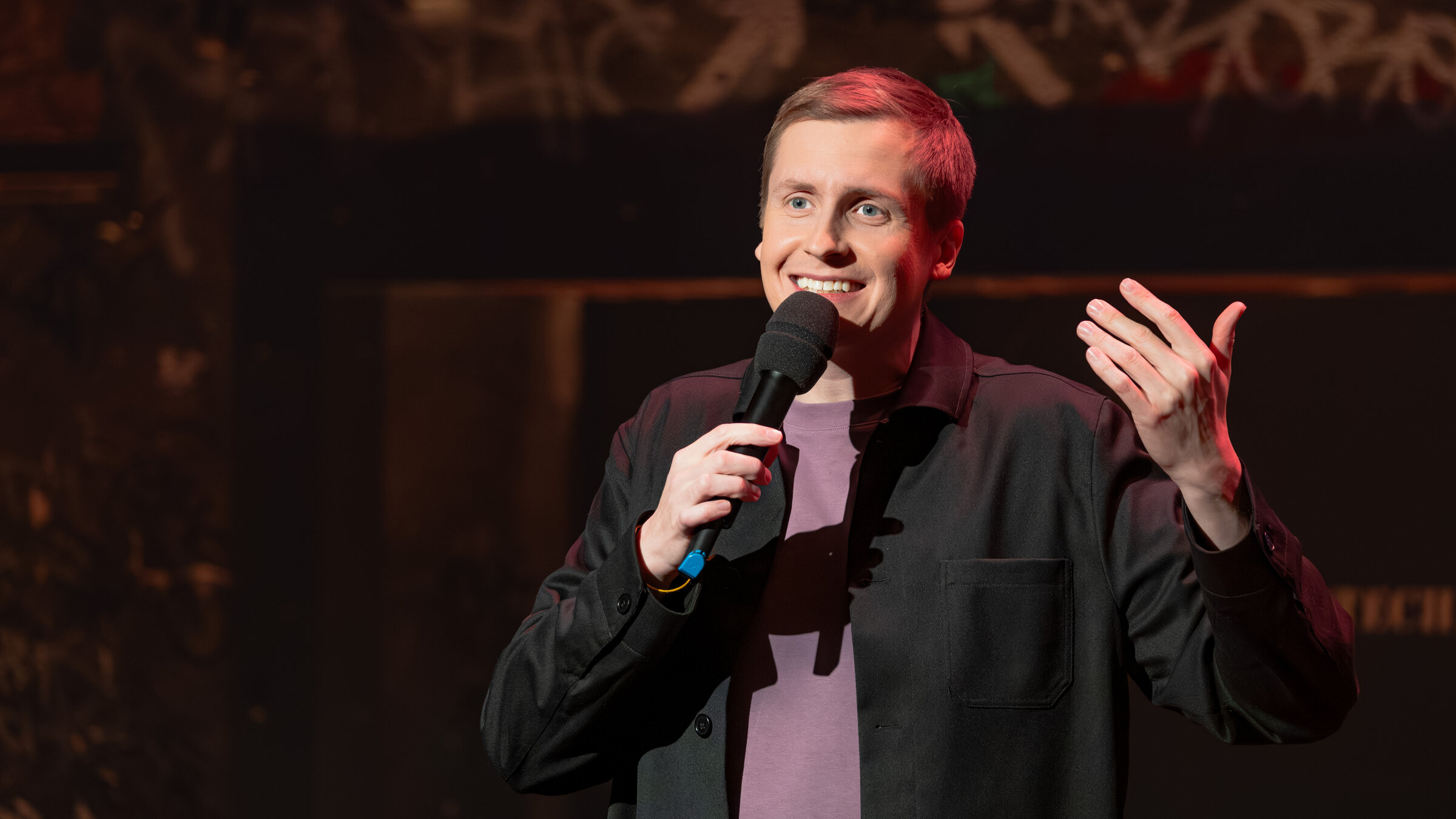 "Till Reiners` Happy Hour": Stand-Up-Comedian und Gastgeber Till Reiners steht mit einem Mikrofon in der Hand auf der Bühne und lächelt Richtung Publikum.
