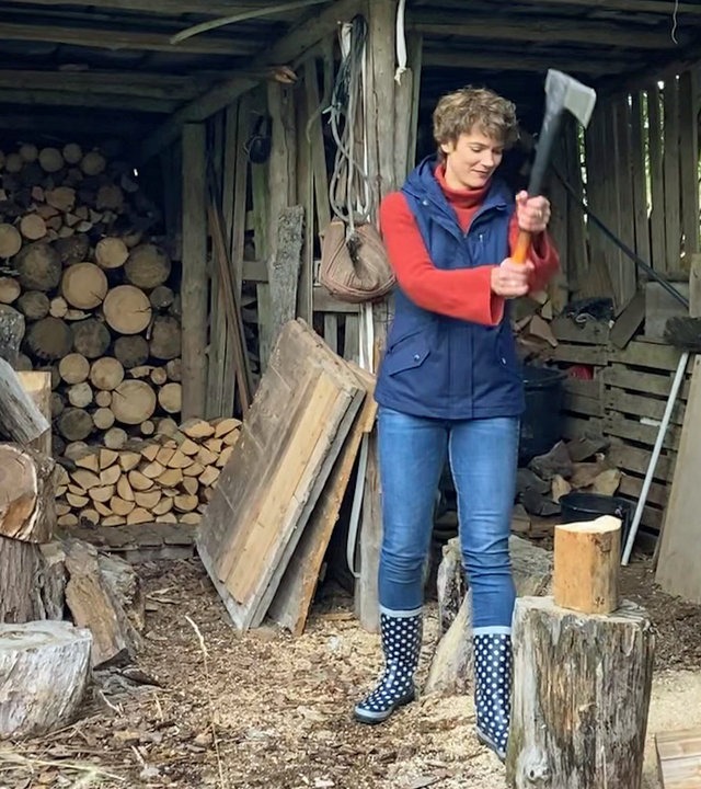 "Terra Xpress: Autarkie: Wege aus der Energiekrise?": Lena Ganschow beim Holzhacken. Sie hebt gerade das Beil. Auf dem Holzklotz liegt ein Stück Holz zum spalten. Neben ihr steht der Besitzer des Selbstversorgungshofes mit verschränkten Händen und schaut ihr zu.