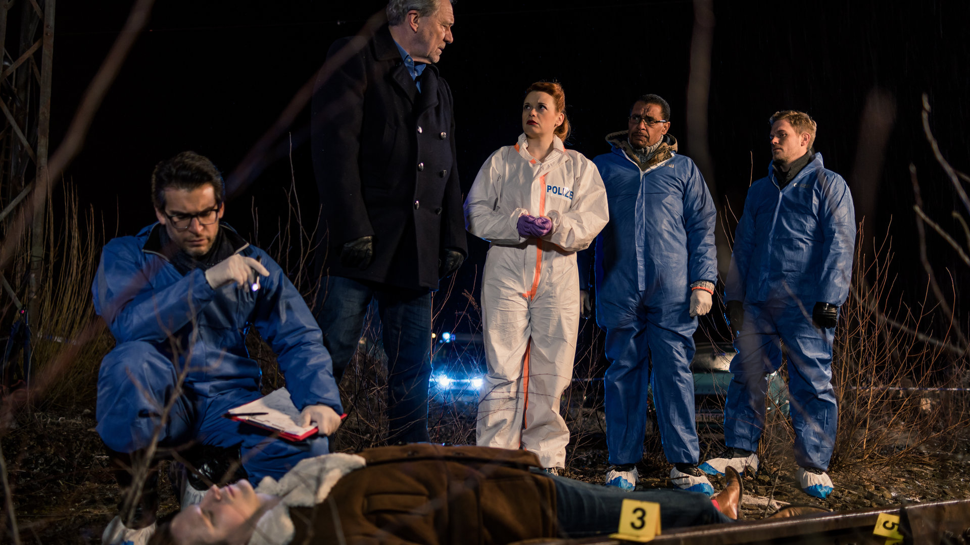 "SOKO München - Fünf Minuten": Am Tatort - Es ist dunkel. Im Hintergrund sind Scheinwerfer eines Polizeiautos zu sehen. Neben einer Gleise liegt eine Leiche auf dem Boden, Dr. Weissenböck (Florian Odendahl) sitzt in der Hocke daneben und ermittelt, er hat eine kleine Taschenlampe in der Hand. Neben ihm stehen Arthur Bauer (Gerd Silberbauer), Billie Curio (Sina Reiß), Franz Ainfachnur (Christofer v. Beau) und Dominik Morgenstern (Joscha Kiefer) und besprechen sich.