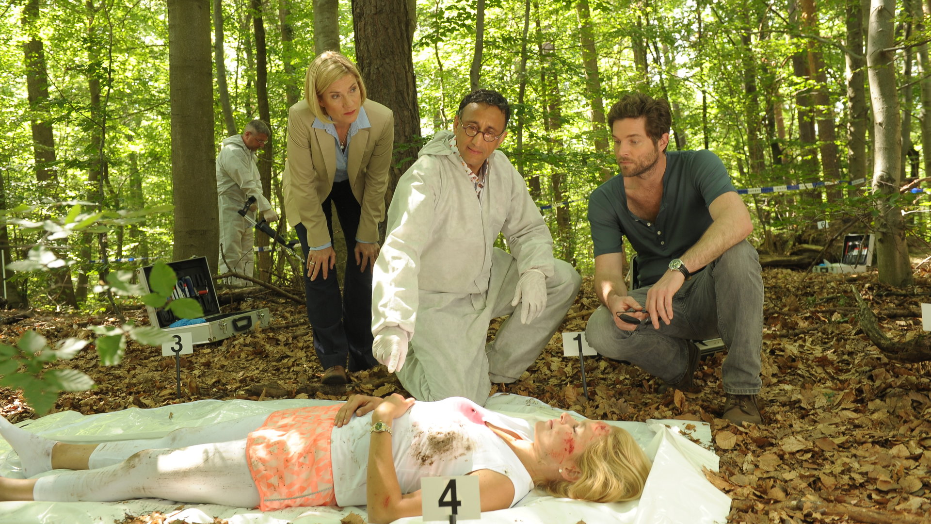 "SOKO Stuttgart - In guten Händen": Martina Seiffert (Astrid M. Fünderich) und Jo Stoll (Peter Ketnath) stehen im Wald beim Opfer Sabine Dombrowski (Komparsin). KTU-Leiter Jan Arnaud (Mike Zaka Sommerfeldt) deutet auf die Hände der Toten, während im Hintergrund ein Spurensicherer (Komparse) den Boden mit einem Metalldetektor absucht.