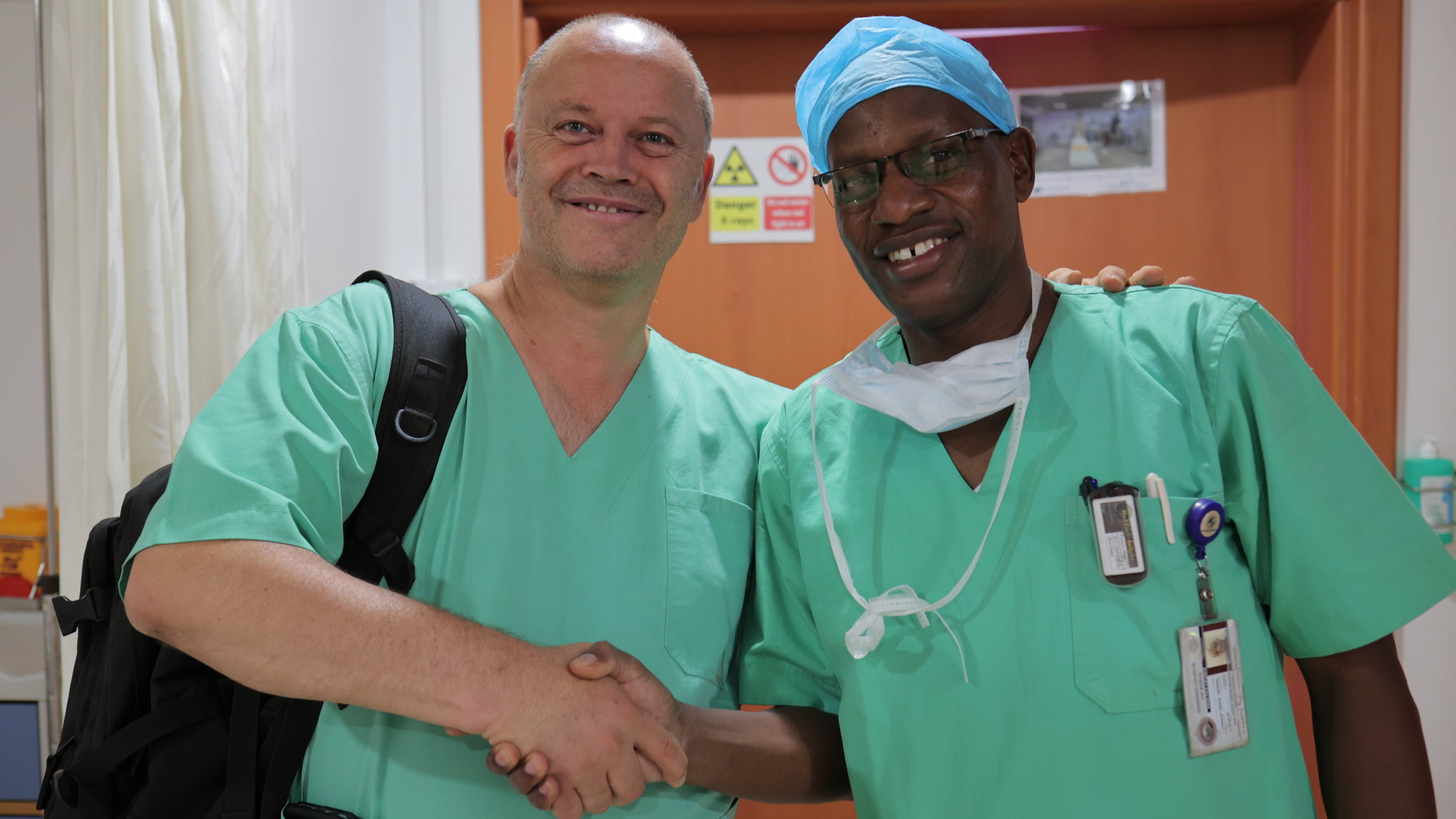 "plan b: Klimaschutz im Krankenhaus - Neue Rezepte für eine nachhaltige Medizin": Dr. Carsten Israel und der kenianische Pfleger Francis stehen in grüner OP-Kleidung auf einem Flur. Sie geben sich die Hand und lächeln in die Kamera.