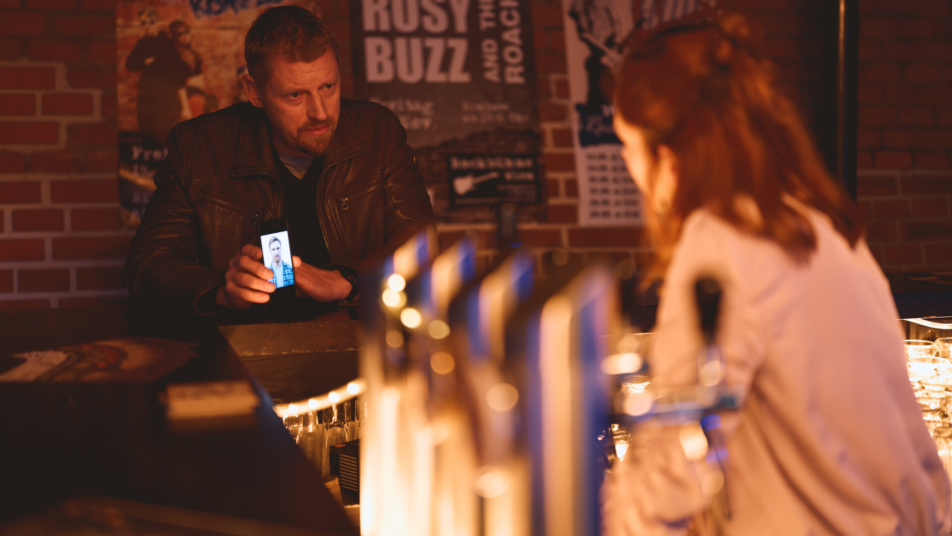 "SOKO Wien - Schlussakkord": Max Herzog (Martin Gruber) sitzt an einer Bar und zeigt Carlotta Benett (Marlena Marie Grübl) das Foto von Günther Herbst auf dem Display eines Smartphones. Hinter Max Herzog hängen einige Konzertplakate an einer roten Backsteinwand.
