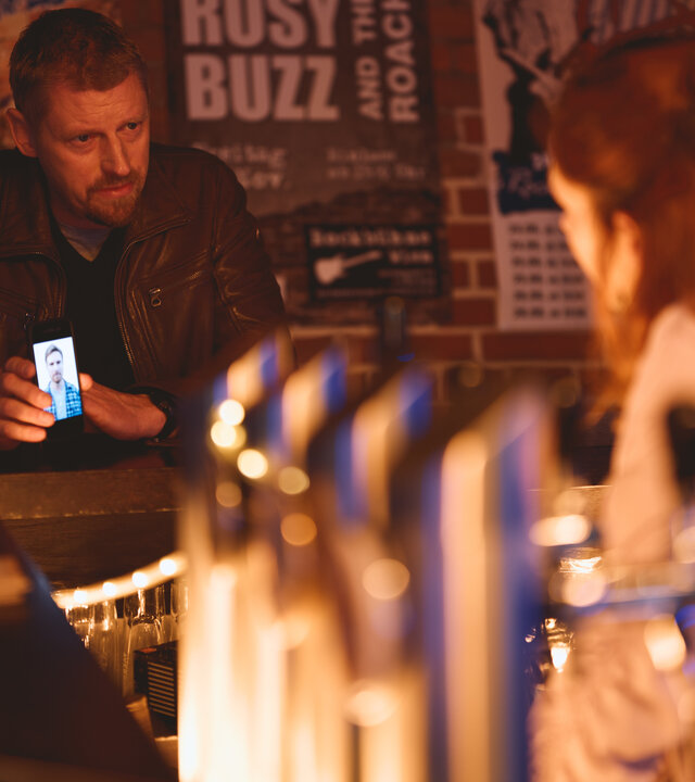 "SOKO Wien - Schlussakkord": Max Herzog (Martin Gruber) sitzt an einer Bar und zeigt Carlotta Benett (Marlena Marie Grübl) das Foto von Günther Herbst auf dem Display eines Smartphones. Hinter Max Herzog hängen einige Konzertplakate an einer roten Backsteinwand.