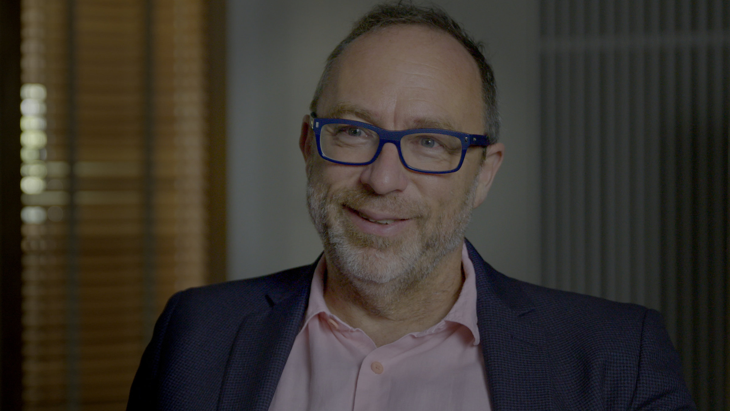 "Wikipedia – Die Schwarmoffensive": Ein Mann mit kurzem grauen Bart und einer Brille mit blauem Rand schaut lächelnd an der Kamera vorbei. Er trägt ein altrosafarbenes Hemd und ein dunkles Sakko.