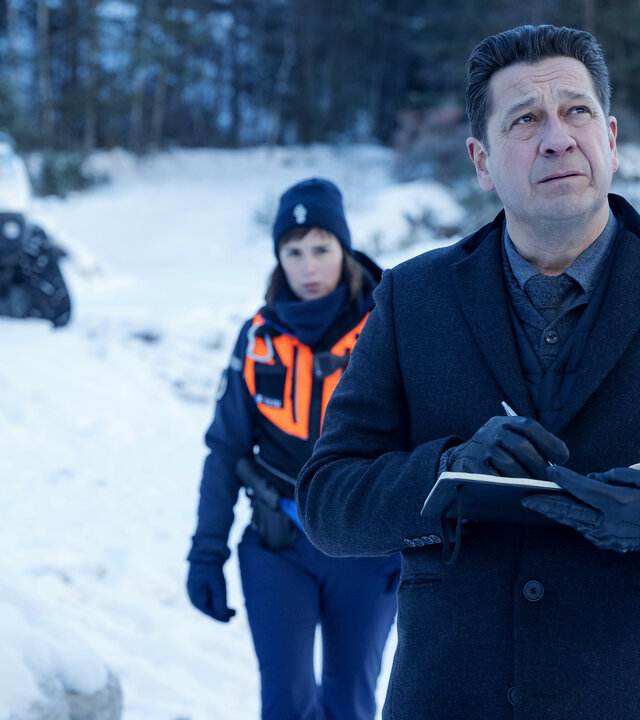 "Tod auf dem Gipfel": Halbnahe: Andréas (Laurent Gerra) steht mit Stift und aufgeschlagenem Notizbuch in einer verschneiten Landschaft und blickt konzentriert nach oben. Hinter Andréas läuft Constance (Clémentine Poidatz) in einer Warnweste auf ihn zu. Im Hintergrund steht ein weißer Jeep am Waldrand geparkt.