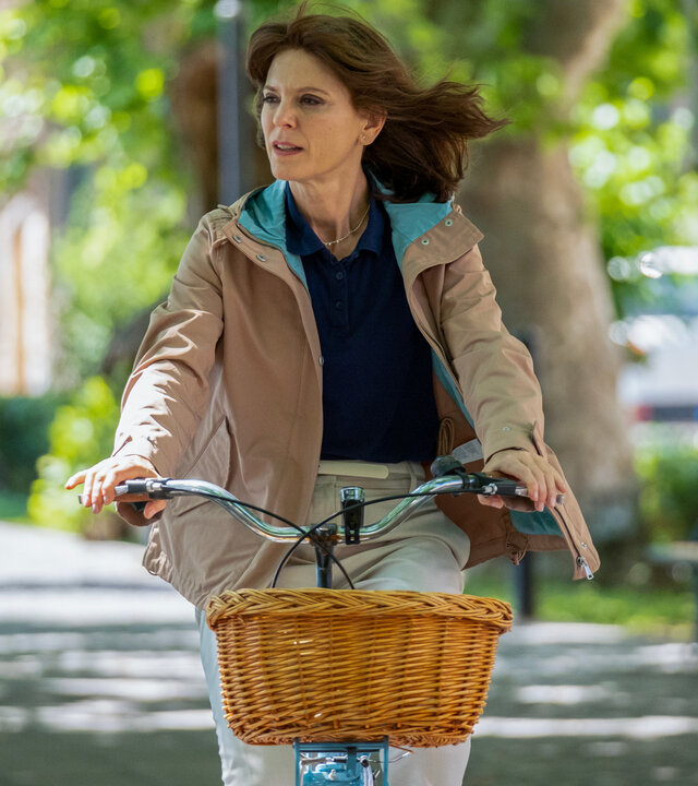 "Signora Volpe - Verstrickung": Sylvia Fox (Emilia Fox) fährt mit einem Fahrrad auf einem von großen Bäumen beschatteten Weg. Sylvia hat sich als "Kirsty" getarnt und trägt eine dunkelhaarige Perücke.