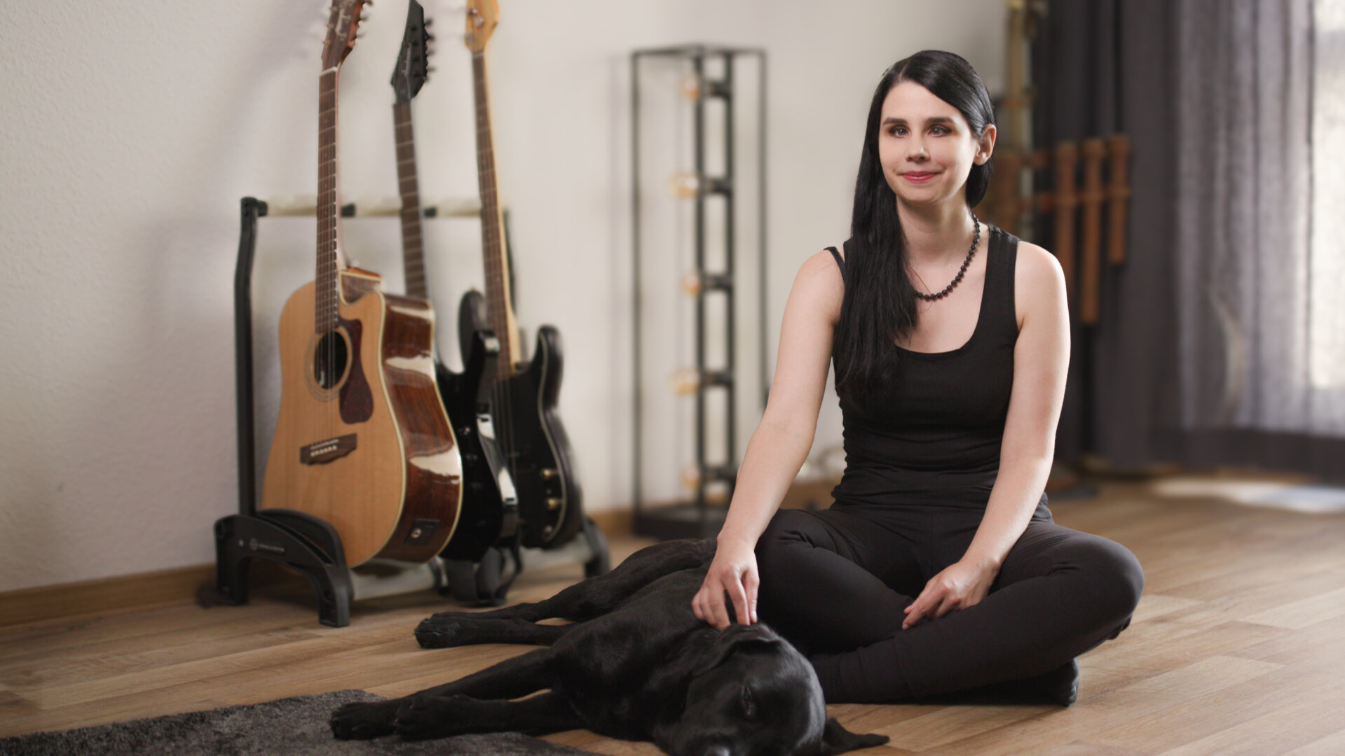 "einfach Mensch - Johanna Kris: Mit Aris durchs Leben": Johanna sitzt im Schneidersitz auf dem Parkett in einem Innenraum. Hinter ihr lehnen drei Gitarren an der Wand, vor ihr liegt ihr Blindenführhund Aris.