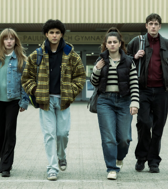 "Von uns wird es keiner sein": Julia (Mina-Giselle Rüffer), Waldi (Lukas von Horbatschewsky), Mina (Derya Akyol) und Tom (Kosmas Schmidt) entfernen sich vom Schuleingang und laufen zusammen über das Schulgelände in Richtung Kamera.