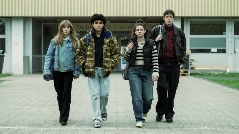 "Von uns wird es keiner sein": Julia (Mina-Giselle Rüffer), Waldi (Lukas von Horbatschewsky), Mina (Derya Akyol) und Tom (Kosmas Schmidt) entfernen sich vom Schuleingang und laufen zusammen über das Schulgelände in Richtung Kamera.