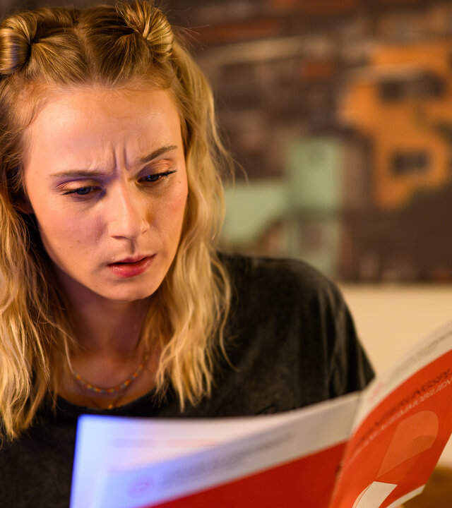 "Bettys Diagnose - Wie ein neuer Mensch": Mia Kunze (Antonia Döring) sitzt auf einer Couch und hält ein Heft in beiden Händen. Im Hintergrund hängt ein Bild an der Wand.