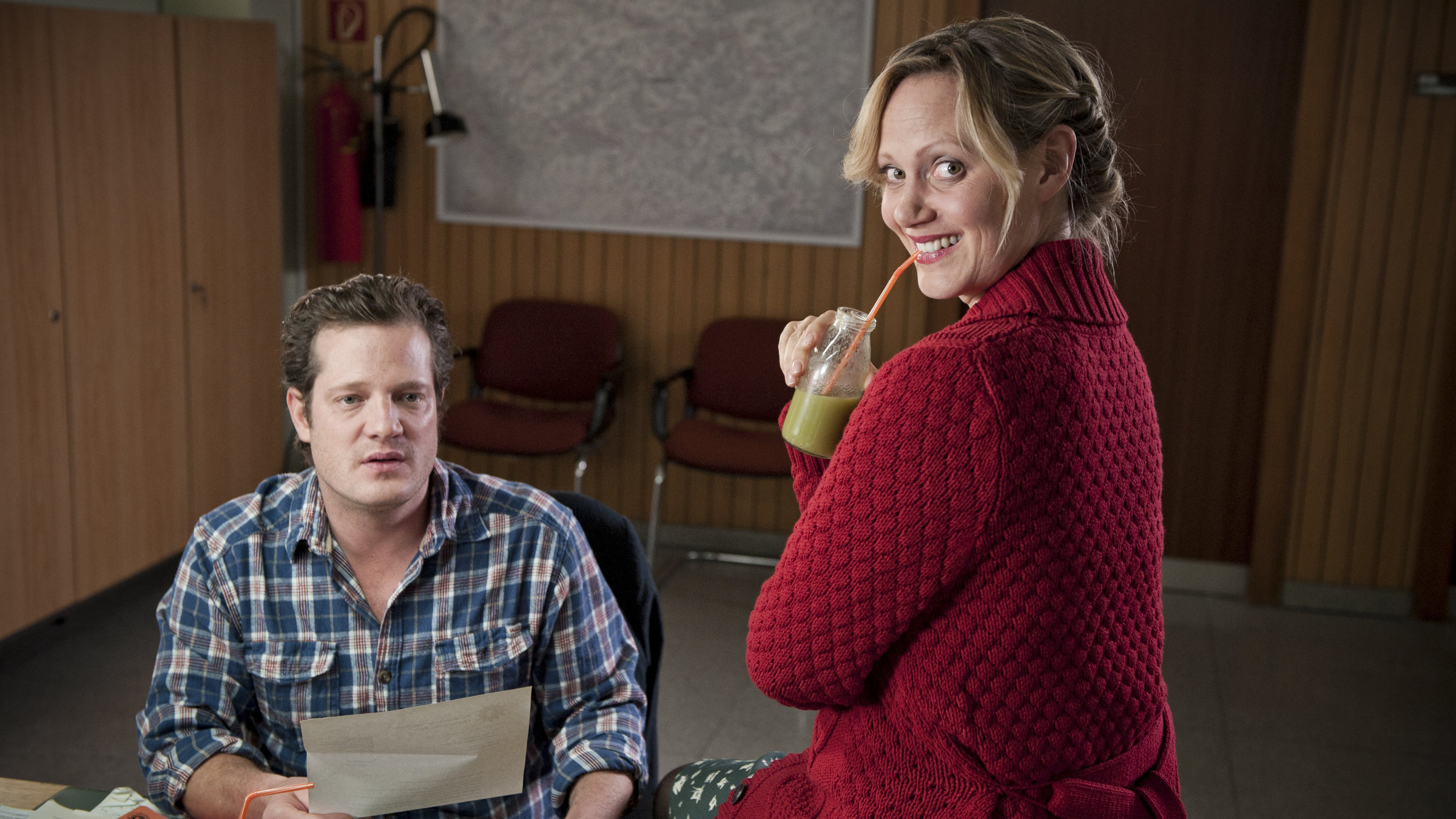 "Mordshunger - Verbrechen und andere Delikatessen: Der Heiler": Britta (Anna Schudt) sitzt auf dem Schreibtisch ihres Bruders Max Janssen (Aurel Manthei) und trinkt einen Saft. Max blättert in seinen Unterlagen.