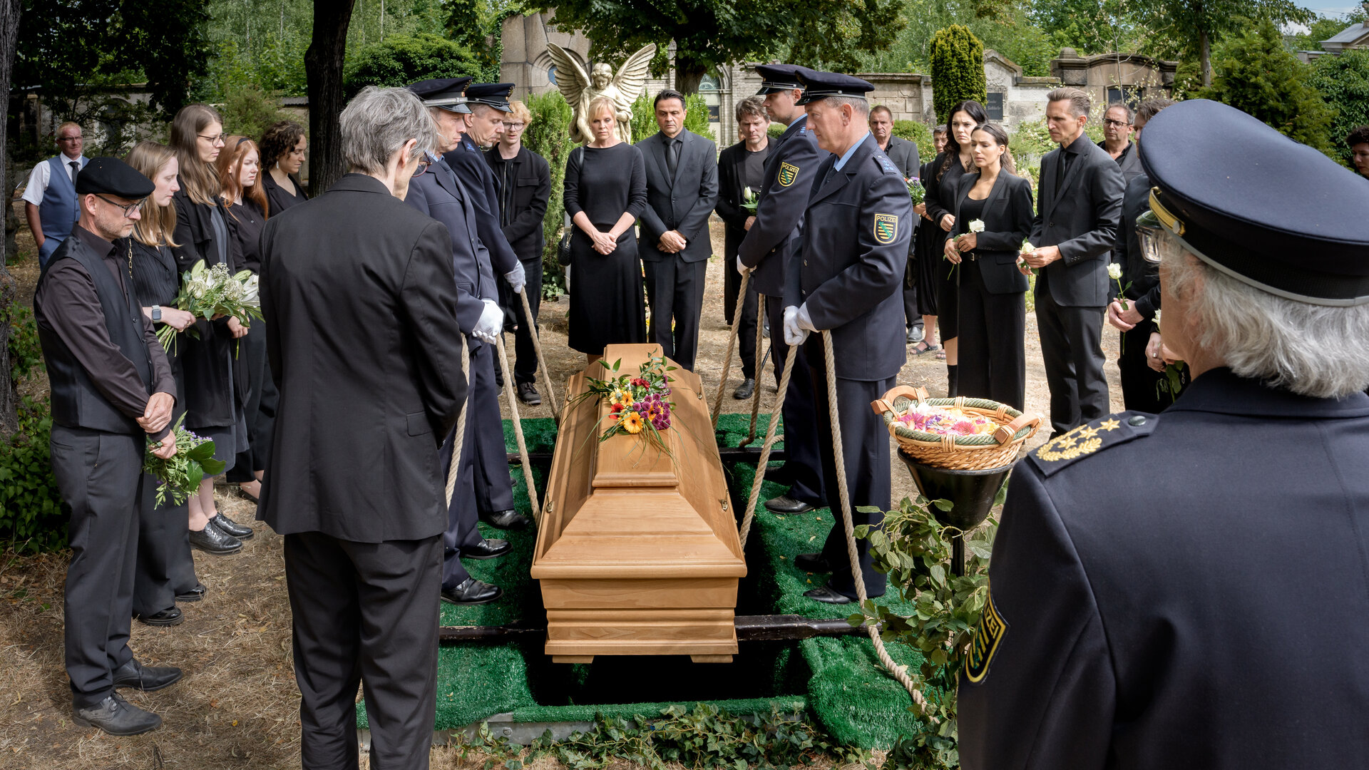 "SOKO Leipzig - Abschied": KTU Holger (Holger Koch), Lorenz Rettig (Daniel Steiner), Paul Zimmermann (Peter Laser), Ina Zimmermann (Melanie Marschke), Staatsanwalt Binz (Michael Rotschopf), Tom Kowalski (Steffen Schroeder), Olivia Fareedi (Nilam Farooq), Kim Nowak (Amy Mußul), Moritz Brenner (Johannes Hendrik Langer) und Komparsen stehen um den Sarg von Jan herum, der gerade beigesetzt wird.