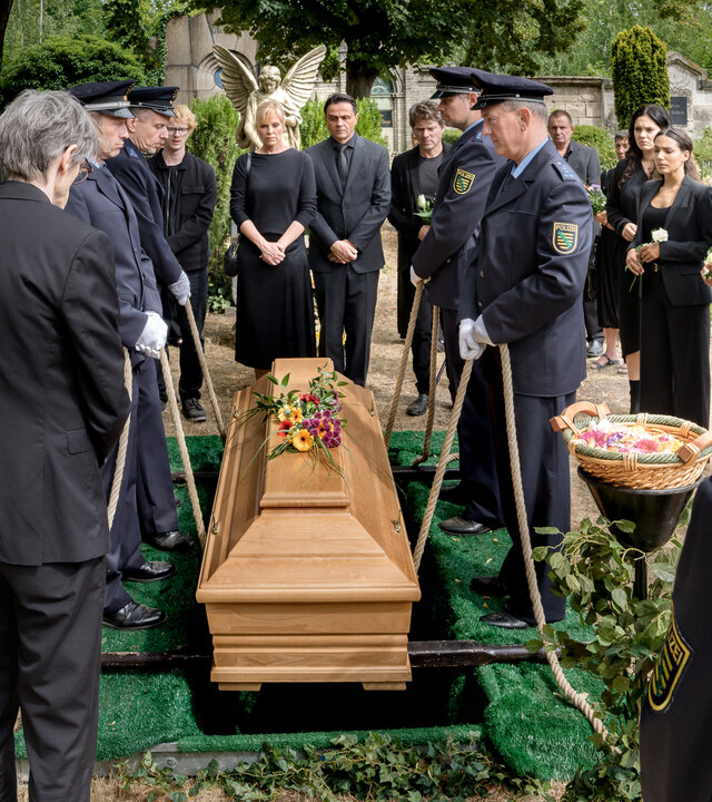 "SOKO Leipzig - Abschied": KTU Holger (Holger Koch), Lorenz Rettig (Daniel Steiner), Paul Zimmermann (Peter Laser), Ina Zimmermann (Melanie Marschke), Staatsanwalt Binz (Michael Rotschopf), Tom Kowalski (Steffen Schroeder), Olivia Fareedi (Nilam Farooq), Kim Nowak (Amy Mußul), Moritz Brenner (Johannes Hendrik Langer) und Komparsen stehen um den Sarg von Jan herum, der gerade beigesetzt wird.