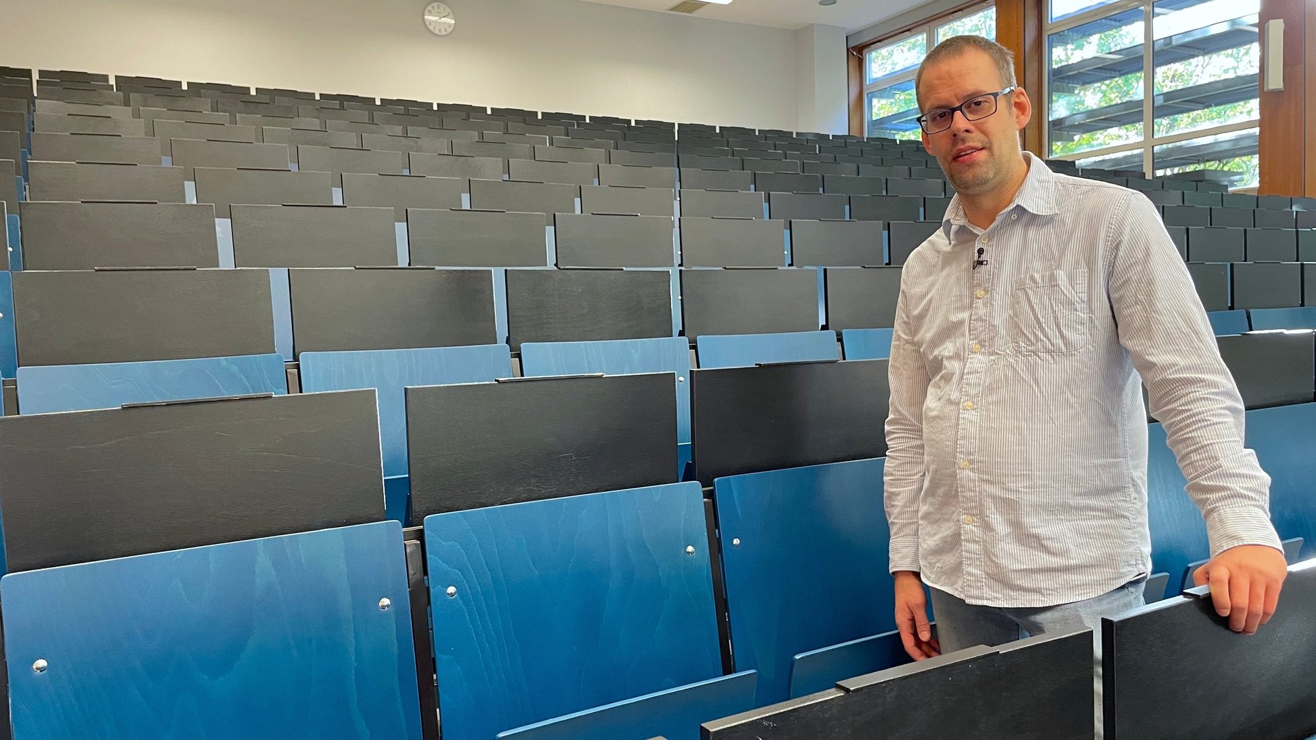 "einfach Mensch: Samuel Wunsch - Von der Werkstatt an die Uni": Samuel Wunsch steht zwischen den Stuhlreihen im Hörsaal. eine Hand liegt auf einer Stuhllehne. Er schaut in die Kamera.