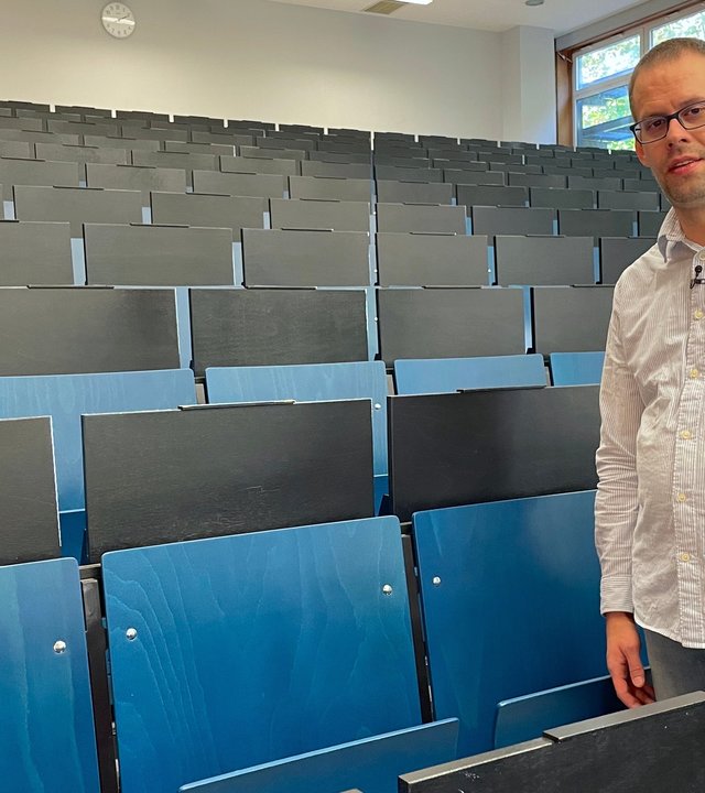 "einfach Mensch: Samuel Wunsch - Von der Werkstatt an die Uni": Samuel Wunsch steht zwischen den Stuhlreihen im Hörsaal. eine Hand liegt auf einer Stuhllehne. Er schaut in die Kamera.