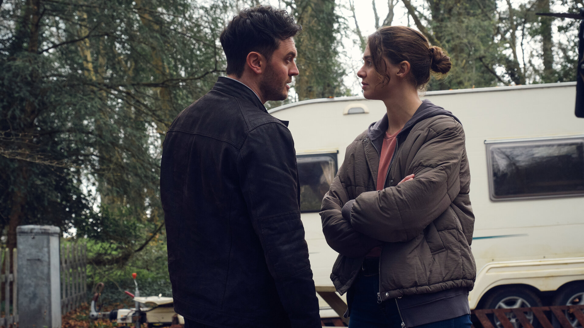 "The Killing Kind: Recht oder Unrecht": Ingrid Lewis (Emma Appleton) steht in einer Daunenjacke mit verschränkten Armen vor DS Nash (Kerr Logan). Die Stimmung ist angespannt, beide schauen sich an. Im Hintergrund steht ein weißer Wohnwagen vor Bäumen.