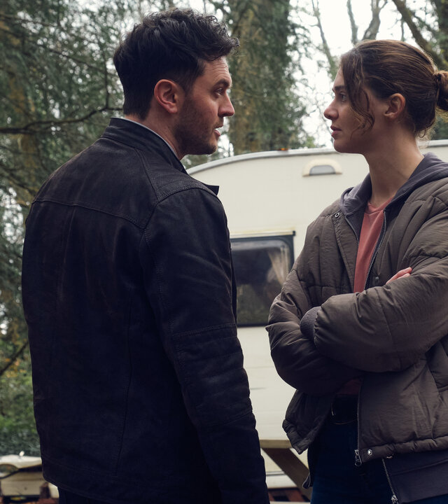 "The Killing Kind: Recht oder Unrecht": Ingrid Lewis (Emma Appleton) steht in einer Daunenjacke mit verschränkten Armen vor DS Nash (Kerr Logan). Die Stimmung ist angespannt, beide schauen sich an. Im Hintergrund steht ein weißer Wohnwagen vor Bäumen.
