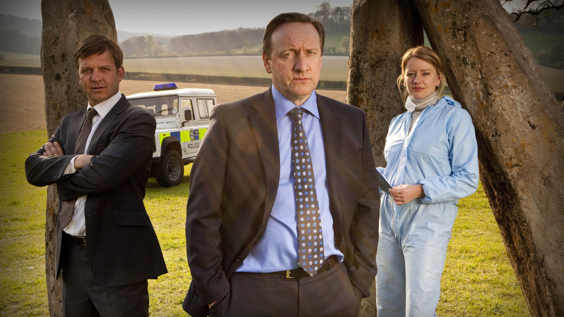"Inspector Barnaby - Die Druiden kommen": DS Ben Jones (Jason Hughes), DCI John Barnaby (Neil Dudgeon), Kate Wilding (Tamzin Malleson) stehen nebeneinander auf einem Feld. Rechts und links von ihnen aufrechtstehende, längliche Steinquader. Alle drei blicken Richtung Kamera. Die Männer tragen Anzüge und Krawatte, Kate Wilding trägt einen hellblauen Schutzanzug. Im Hintergrund ein weißer Polizeijeep.