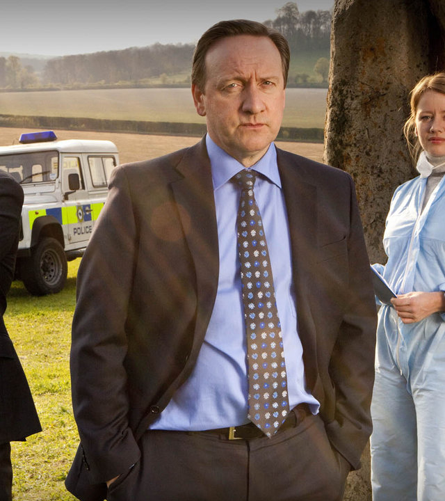 "Inspector Barnaby - Die Druiden kommen": DS Ben Jones (Jason Hughes), DCI John Barnaby (Neil Dudgeon), Kate Wilding (Tamzin Malleson) stehen nebeneinander auf einem Feld. Rechts und links von ihnen aufrechtstehende, längliche Steinquader. Alle drei blicken Richtung Kamera. Die Männer tragen Anzüge und Krawatte, Kate Wilding trägt einen hellblauen Schutzanzug. Im Hintergrund ein weißer Polizeijeep.