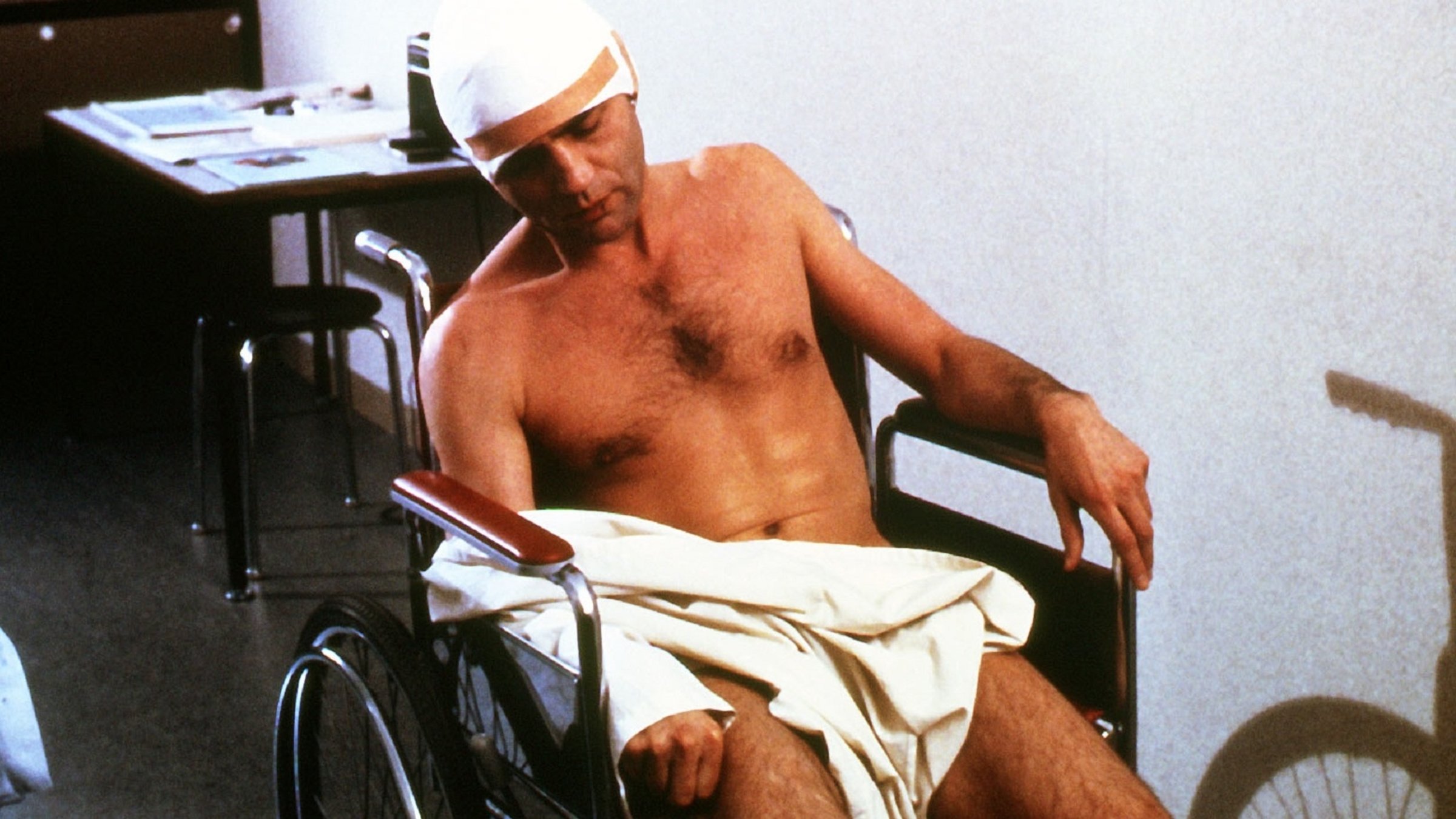 "Messer im Kopf": Ein nackter Mann (Bruno Ganz) sitzt mit bandagiertem Kopf und einem Kittel über dem Schoß in einem Krankenhausrollstuhl.