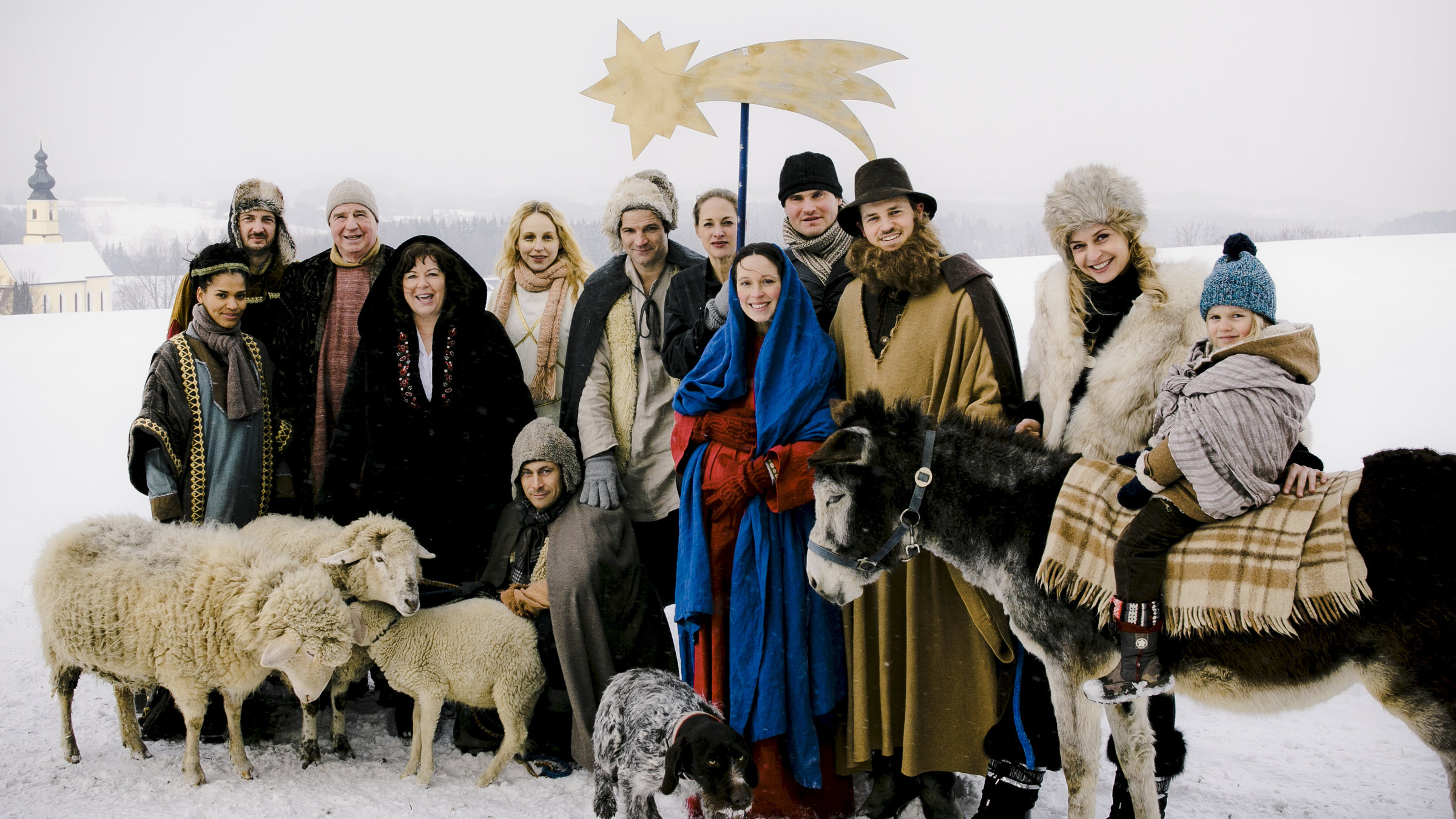 "Beste Bescherung": Malia (Cynthia Micas), Aimer (Jan Messutat), Karl (Friedrich von Thun), Rosalie (Gundi Ellert), Susan (Petra Schmidt-Schaller), Ben (Misel Maticevic), Anna (Sophie von Kessel), Miriam (Anneke Schwabe), Tom (Fabian Hinrichs), Stefan (Frederic Linkemann), Dina (Franziska Schlattner), Sharif (Arian Montgomery) und stehen in einer Gruppe zusammen, davor sitzt Vince (Marc Hosemann) in einer verschneiten Landschaft. Vor der Gruppe stehen Schafe, ein Hund und ein Esel, auf dem Sharif (Arian Montgomery) sitzt. In der Mitte hält jemand einen großen Stern an einem Stab hoch.