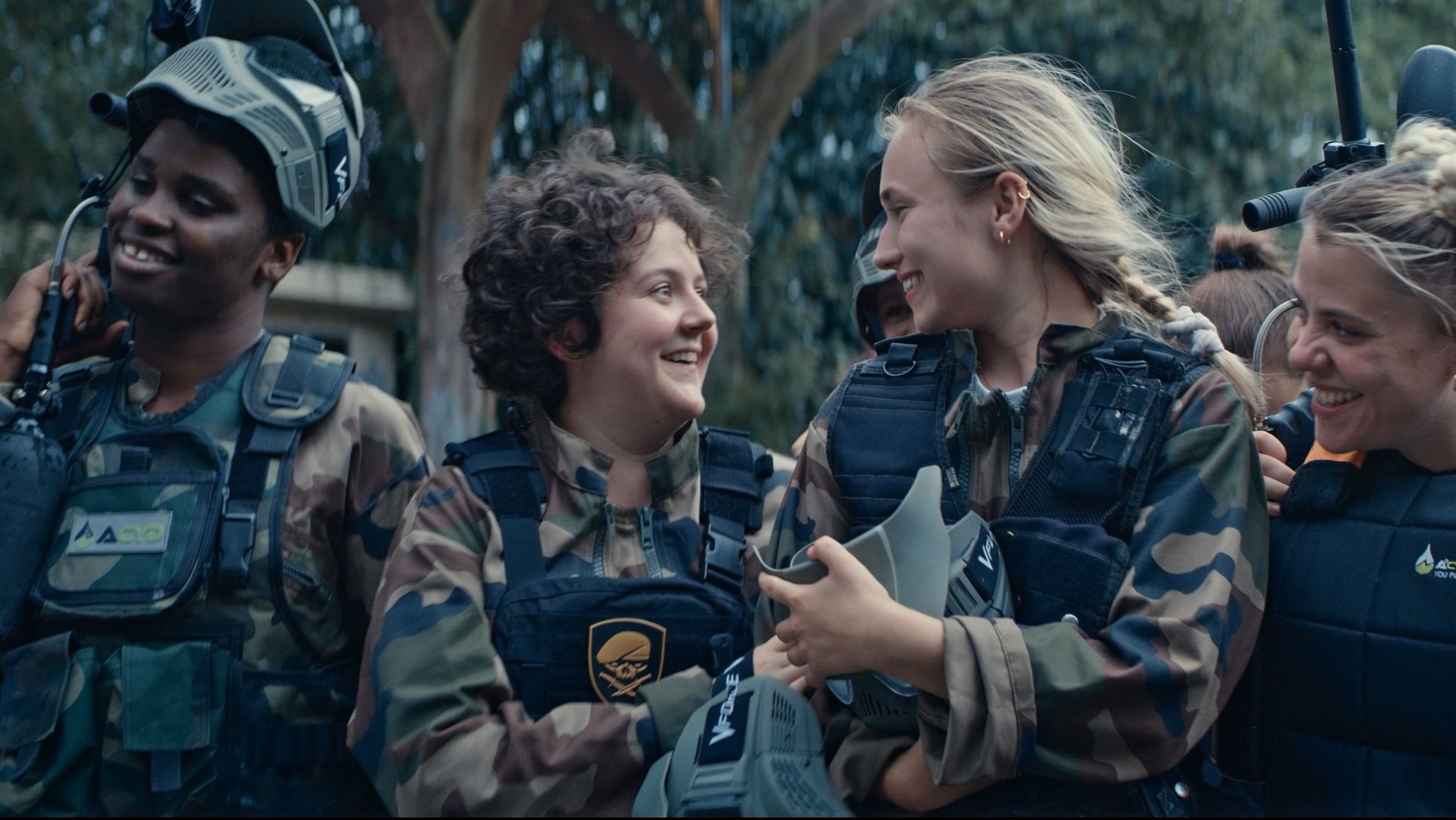 "Derby Girl - Scharf kalkuliert": Acid Cyprine (Jisca Kalvanda), Marie-Josée (Salomé Diénis Meulien), Lola Bouvier (Chloé Jouannet), Mother Blocker (Sophie-Marie Larrouy), Harry Pod''Couilles (Yilin Yang) und Booby Mercury (Allison Chassagne) laufen lachend nebeneinander im Wald. Alle tragen Armee-Klamotten, Sicherheitswesten und Helme.