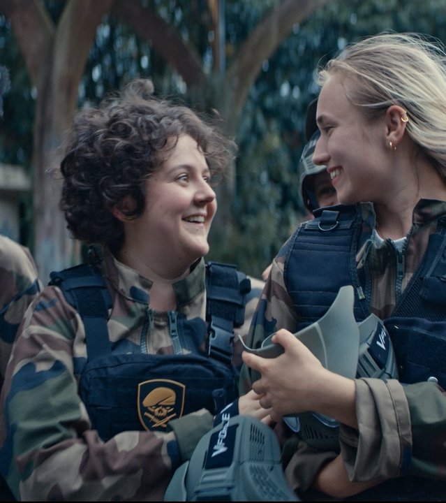 "Derby Girl - Scharf kalkuliert": Acid Cyprine (Jisca Kalvanda), Marie-Josée (Salomé Diénis Meulien), Lola Bouvier (Chloé Jouannet), Mother Blocker (Sophie-Marie Larrouy), Harry Pod''Couilles (Yilin Yang) und Booby Mercury (Allison Chassagne) laufen lachend nebeneinander im Wald. Alle tragen Armee-Klamotten, Sicherheitswesten und Helme.