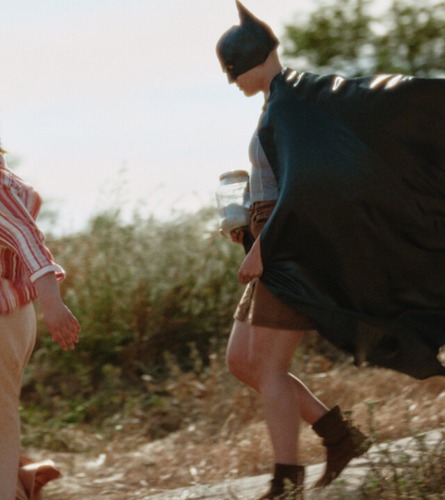 "How to Kill Your Sister - Folge 6": Kat (Marjan De Schutter) und Anna (Emma Rotsaert) gehen einen Abhang herunter. Im Hintergrund sind unscharf Bäume und Büsche zu erkennen. Die Sonne scheint. Anna trägt eine Batman-Maske mitsamt Umhang, Kat eine Pferdemaske.