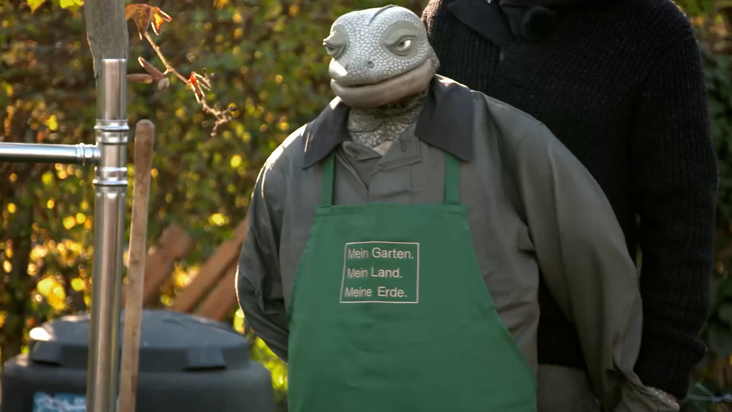 "Die Echse auf Achse - Im Schrebergarten": Die Echse steht mit einer grünen Gärtnerschürze im Garten.
