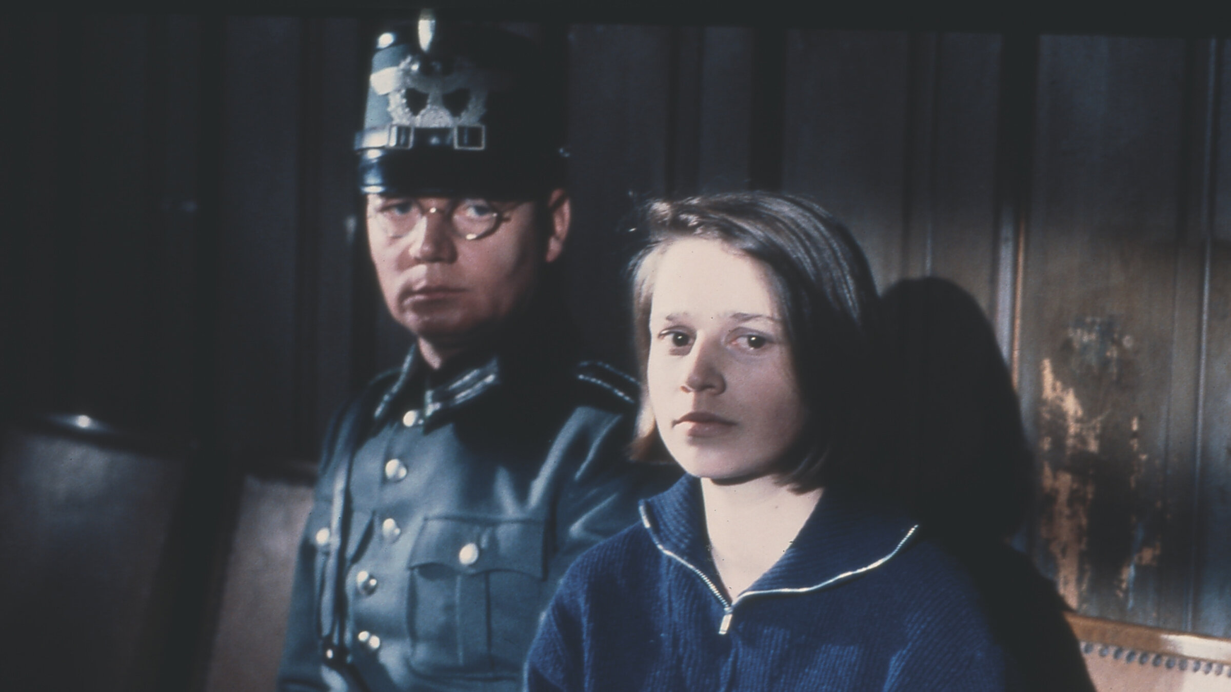 "Die weiÃe Rose": Sophie Scholl (Lena Stolze) trÃ¤gt einen blauen Troyer und sitzt mit traurigem Blick neben einem Polizisten (Darsteller unbekannt) vor einer abgewetzten holzvertÃ¤felten Wand.